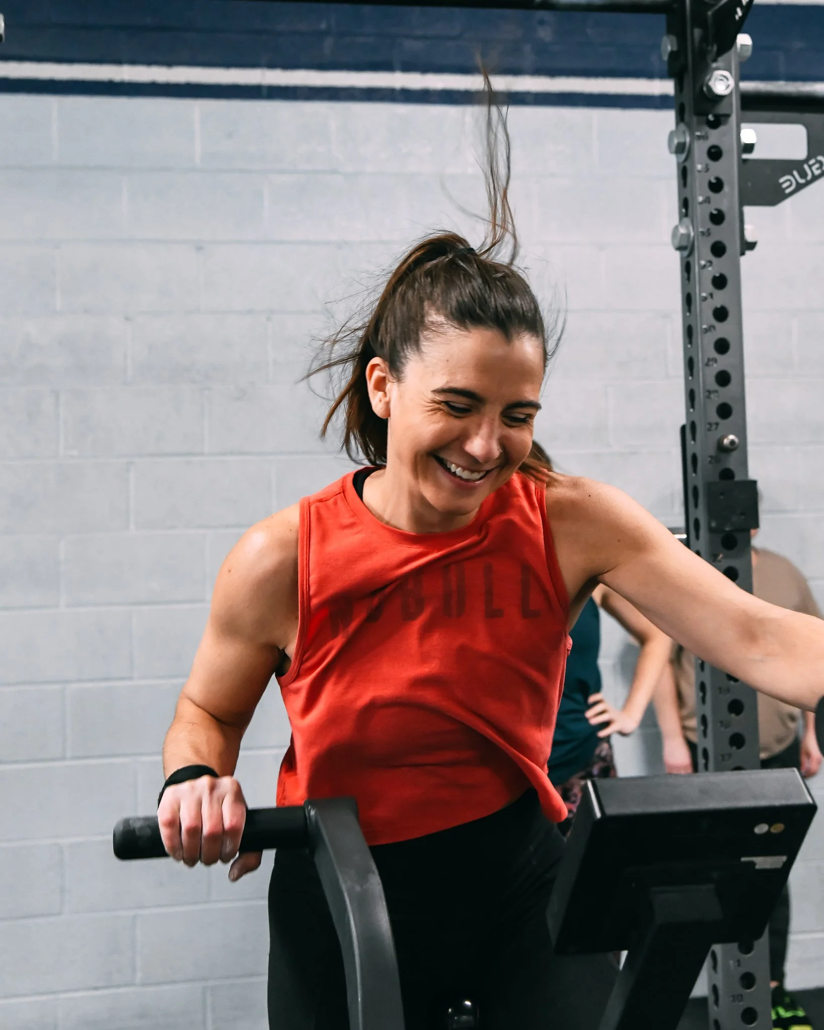 A woman with brown hair in a ponytail smiling as she works out with a barbell in a gym. She is wearing a red sleeveless shirt and black pants, with a gray wall in the background.