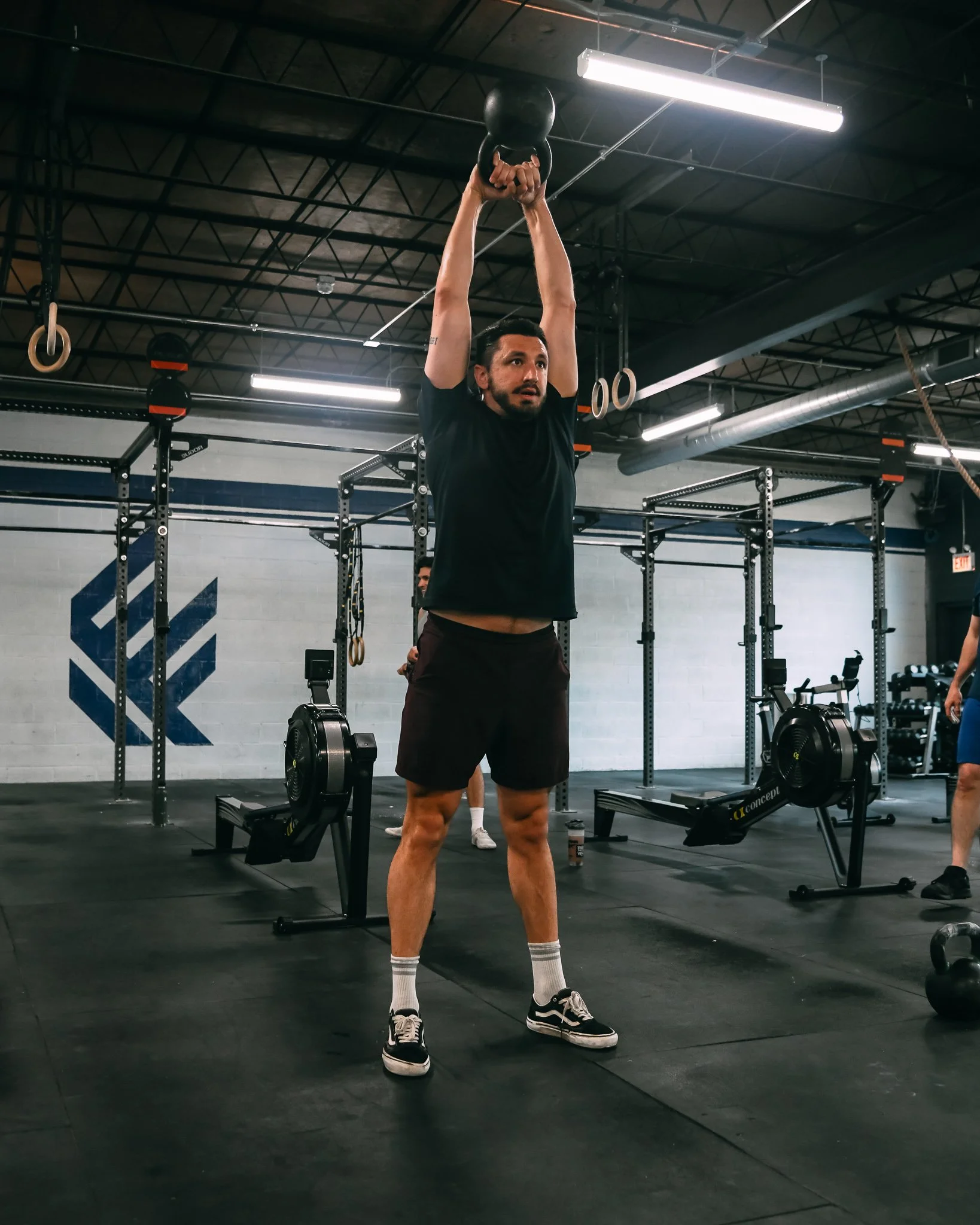 A man in a black t-shirt, black shorts, white socks, and sneakers is lifting a kettlebell with both hands overhead in a gym. The gym has black floors, metal racks, workout equipment, and gymnastic rings hanging from the ceiling. The lighting is fluorescent, and there are other people visible in the background.