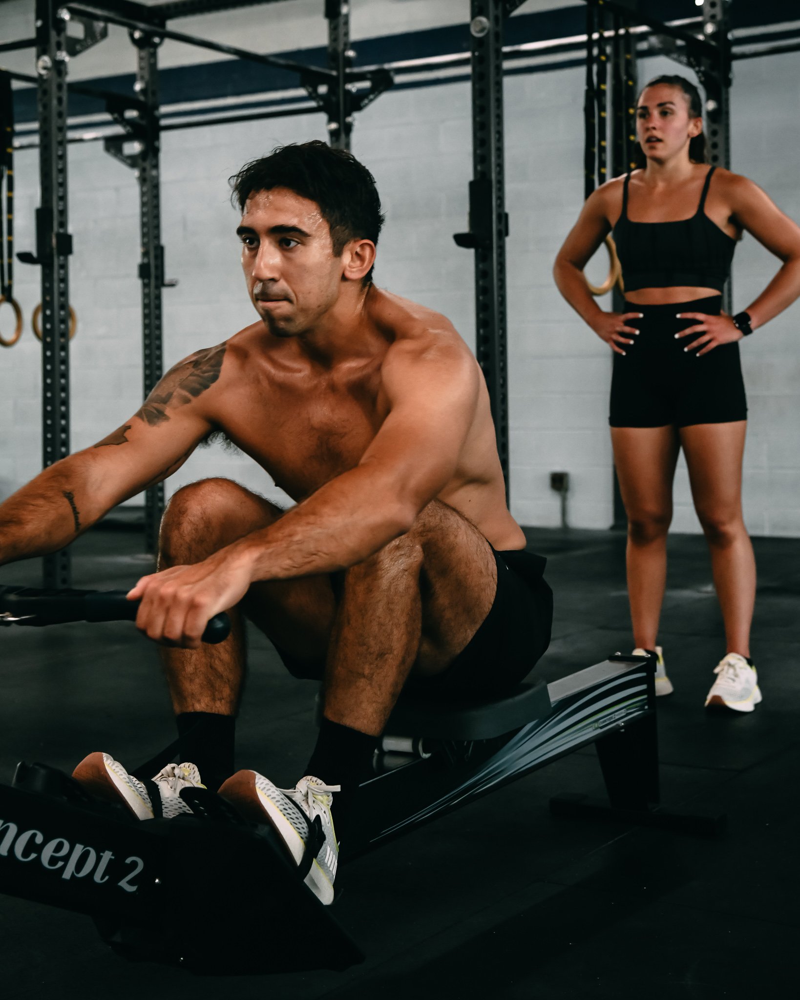 A man with black hair and a tattoo on his left arm, shirtless, is sitting on a rowing machine in a gym, exerting effort with sweat visible on his face. Behind him, a woman with long dark hair tied back, wearing a black sports bra and shorts, stands with her hands on her hips, watching him. The gym has a gray brick wall and black workout equipment, including pull-up bars and gymnastic rings.