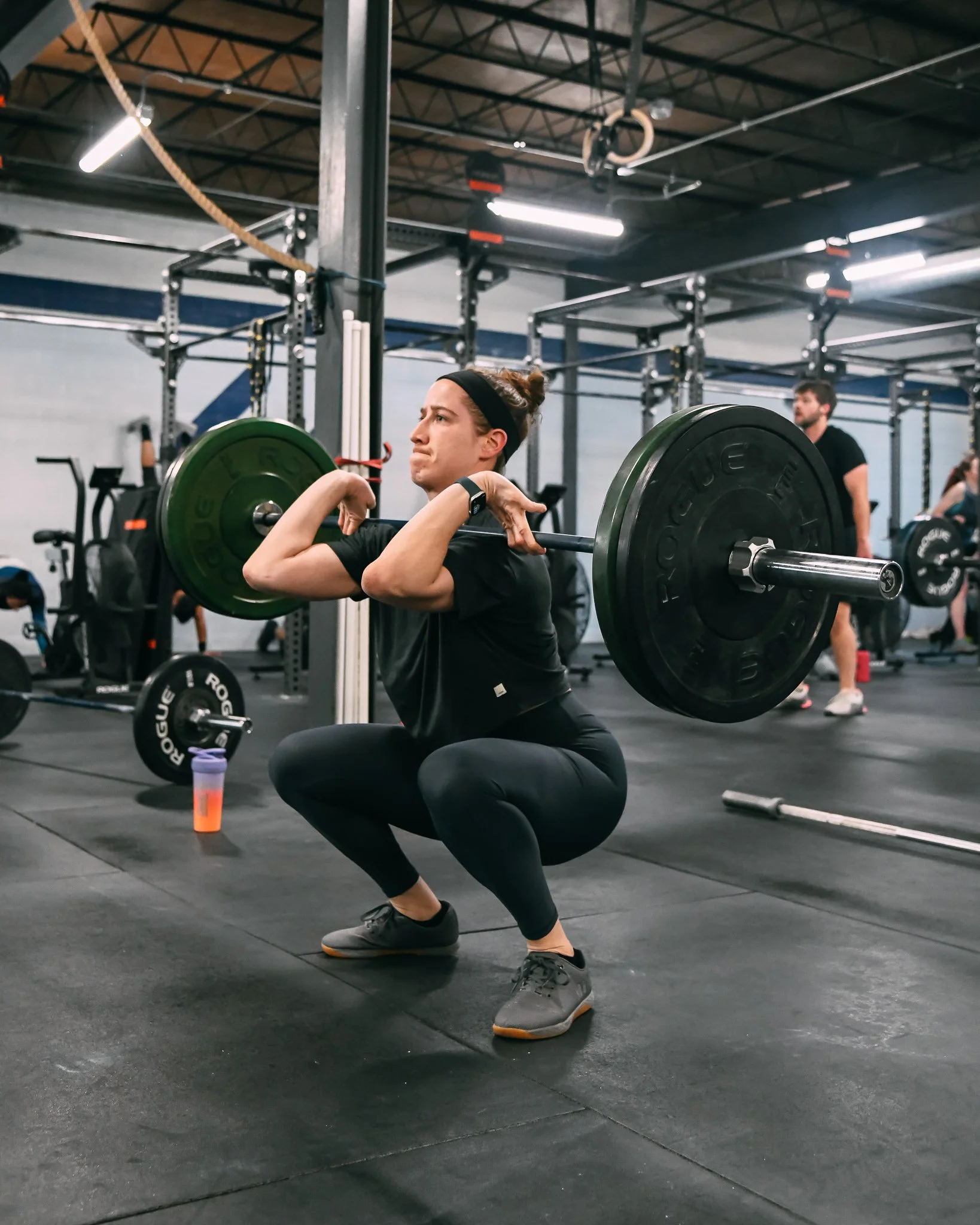A woman performing a squat exercise with a barbell loaded with weight plates at Feast CrossFit near Roscoe Village. She is wearing black workout clothes, gray sneakers, and a headband, with a focused expression.