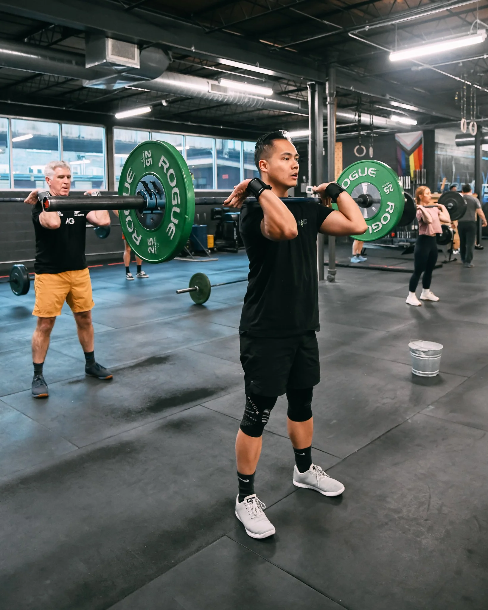 Two men lifting barbells with green weight plates in a gym, with others working out in the background.
