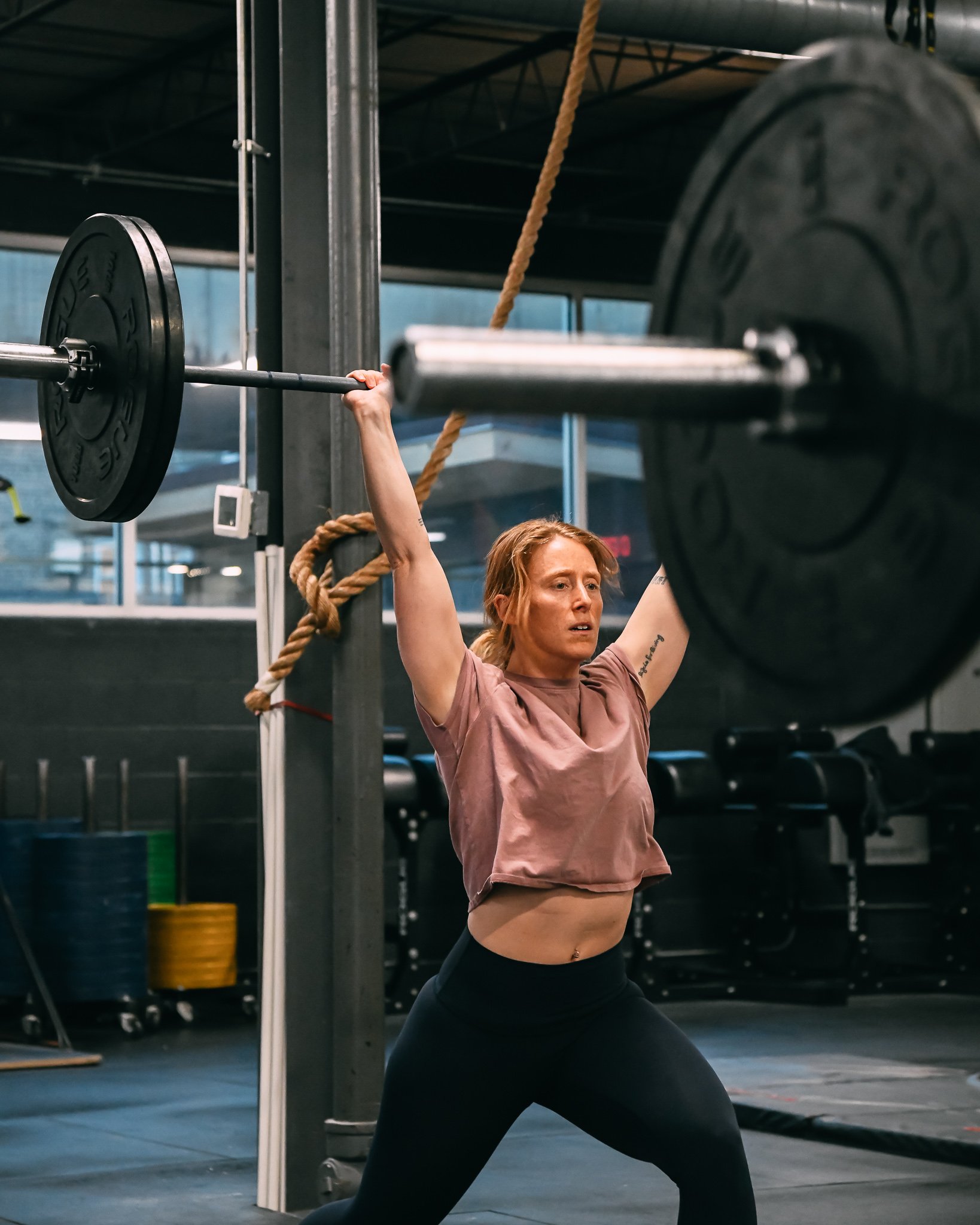 A woman performs an overhead barbell shoulder press in a gym, with a large weight plate on each side of the barbell, and gym equipment visible in the background.