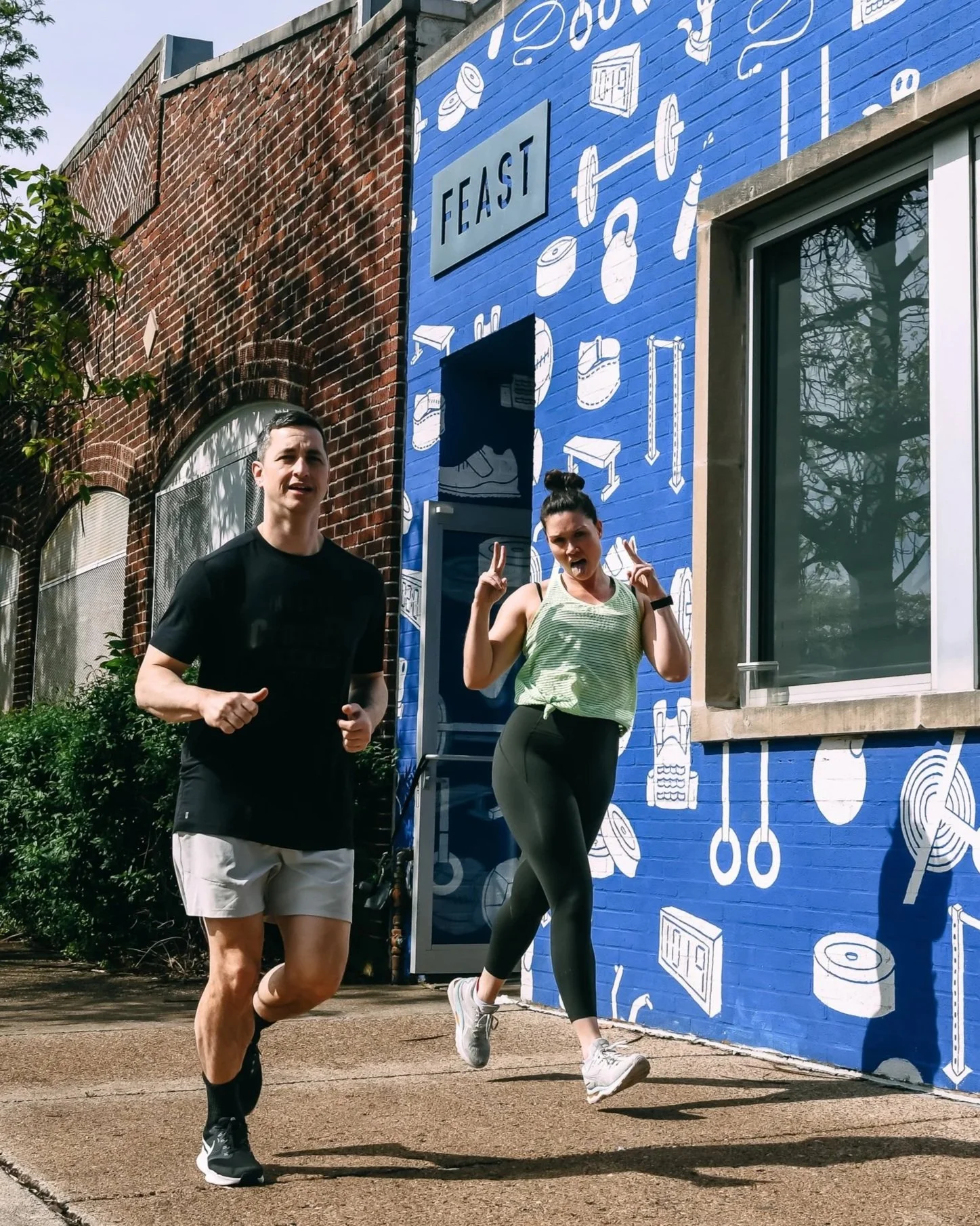 Two people jogging outdoors in front of a blue wall with white illustrations of food and kitchen items, and a blue sign with the word "EAST" on it.