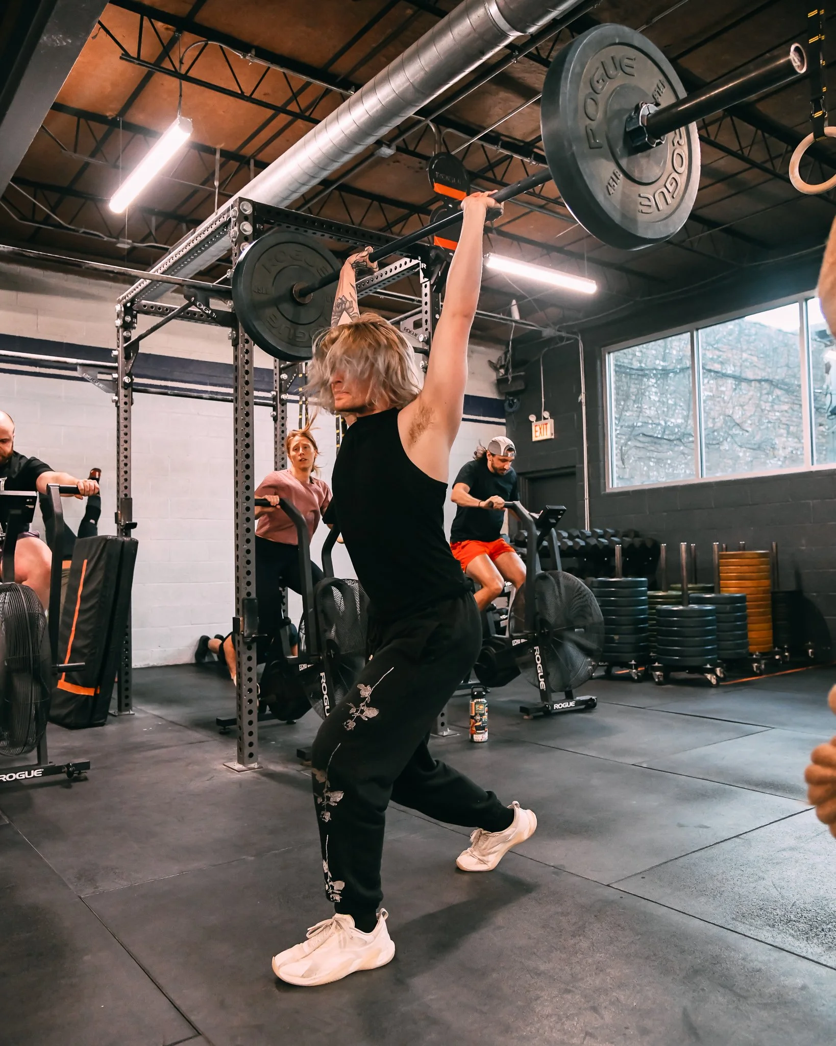 A woman with blonde hair, dressed in black athletic wear and white sneakers, is performing a kettlebell exercise in a gym. She is holding a kettlebell above her head with both hands, surrounded by other gym members, weights, and exercise equipment.