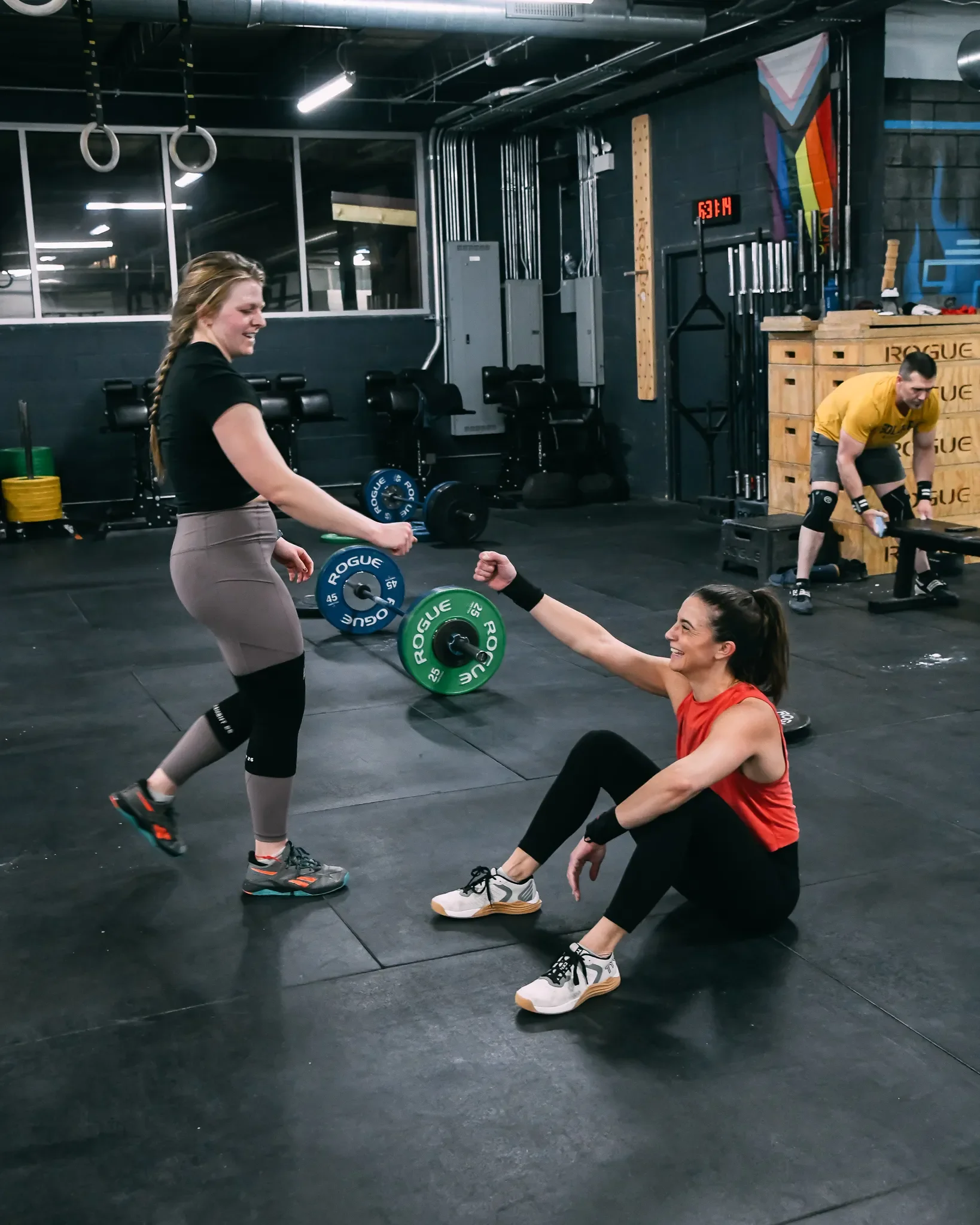 Two female athletes smiling and fist-bumping with a barbell on the floor in the background after a workout at Feast Fitness