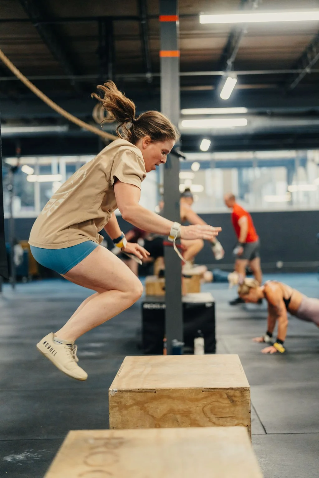 A woman jumping onto a wooden box during a workout session at a gym, with other people exercising in the background.