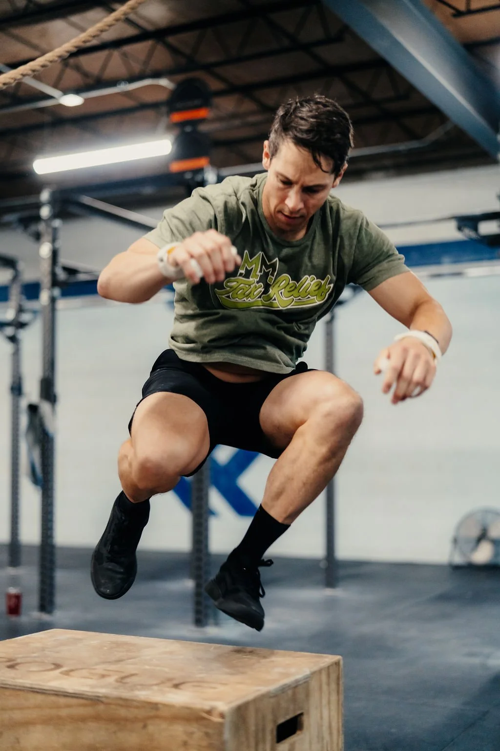 A man in an athletic shirt and shorts is mid-air performing a box jump at a gym. He is about to land on a wooden box during a workout session.