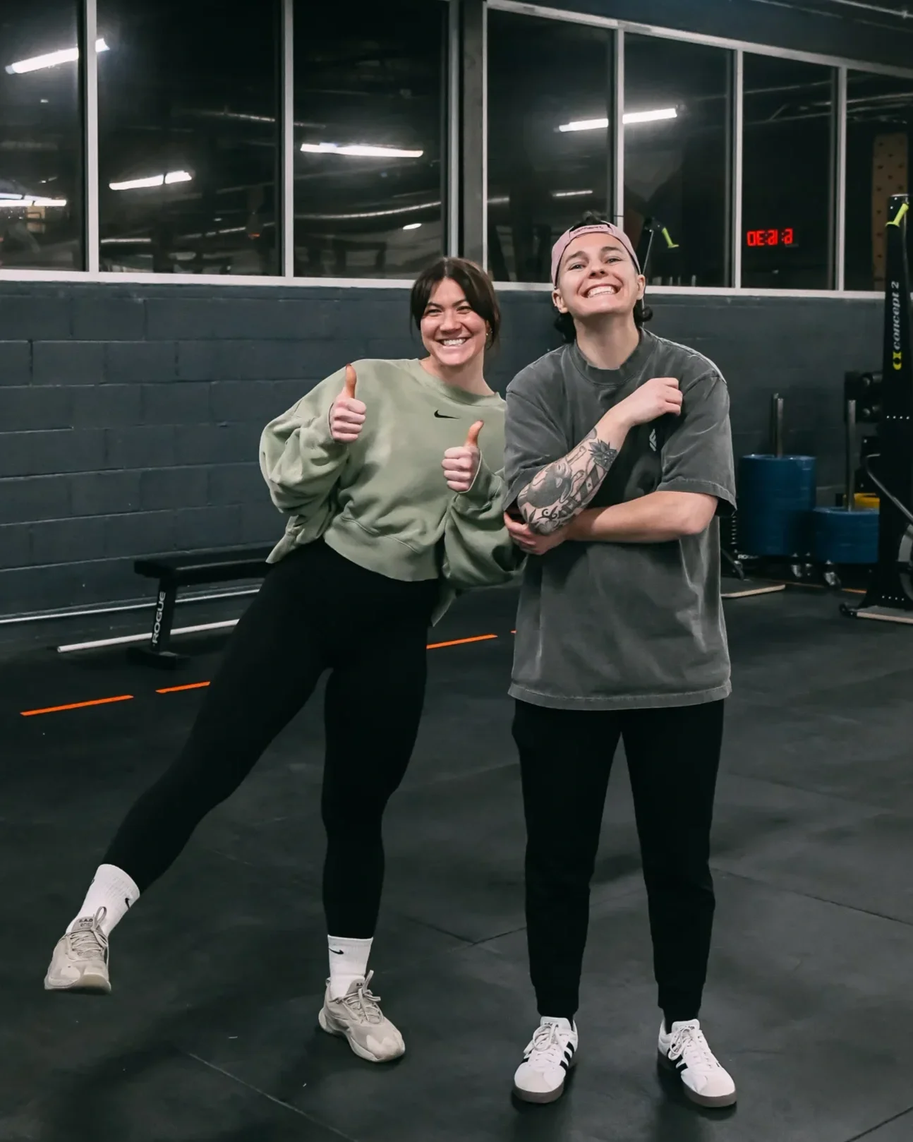 Two women smiling and posing in a gym, one giving a thumbs up and the other with arms crossed, with fitness equipment and a digital clock in the background.