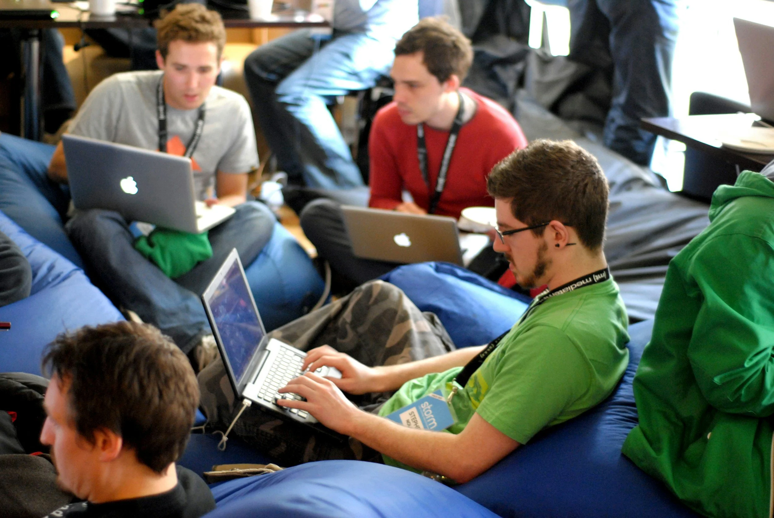 Group of young people sitting together on bean bags in a classroom, using laptops and engaging in discussion.