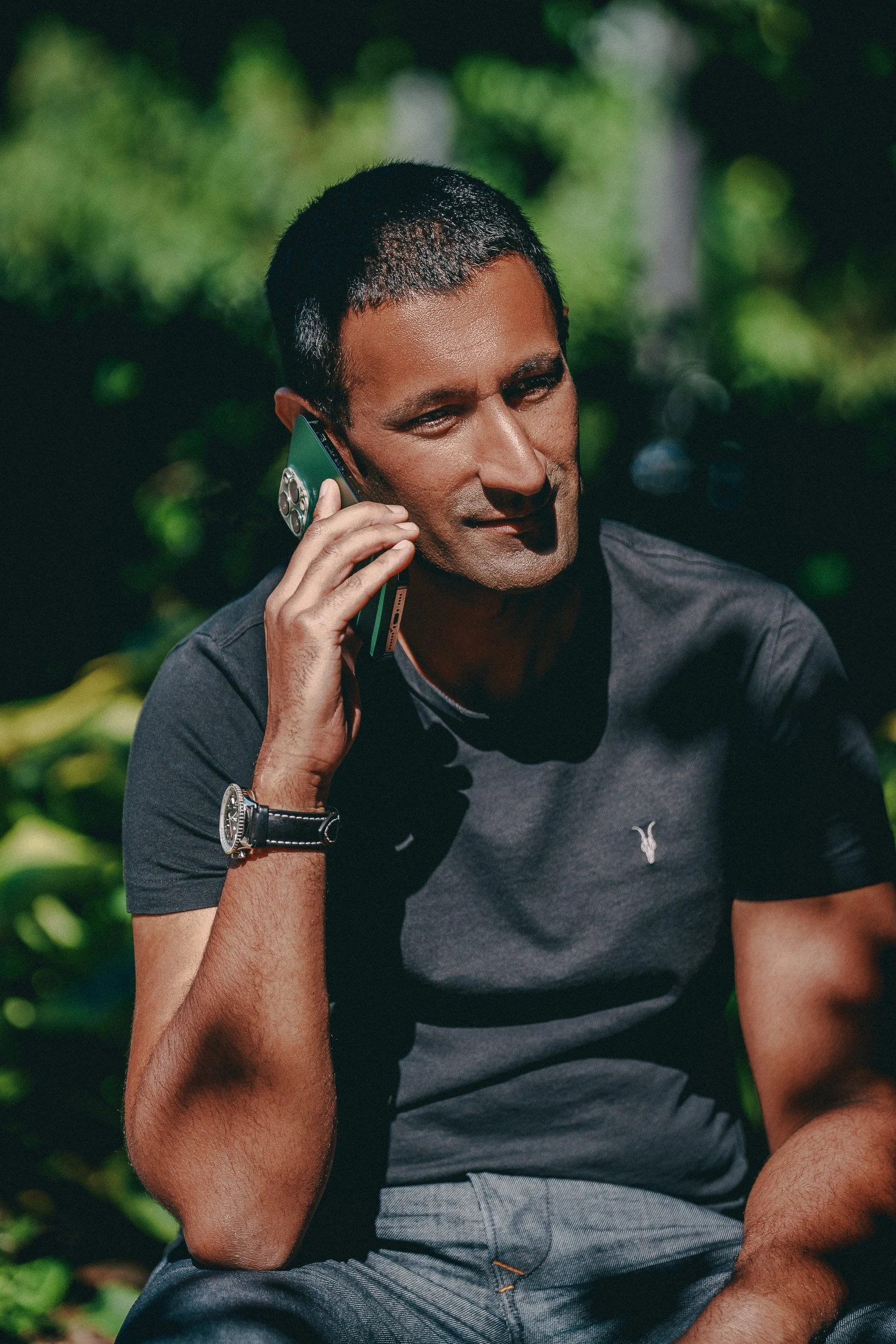 Dr Milan Mehta with short black hair talking on a cellphone outdoors, wearing a black t-shirt and a watch, with a blurred green leafy background.
