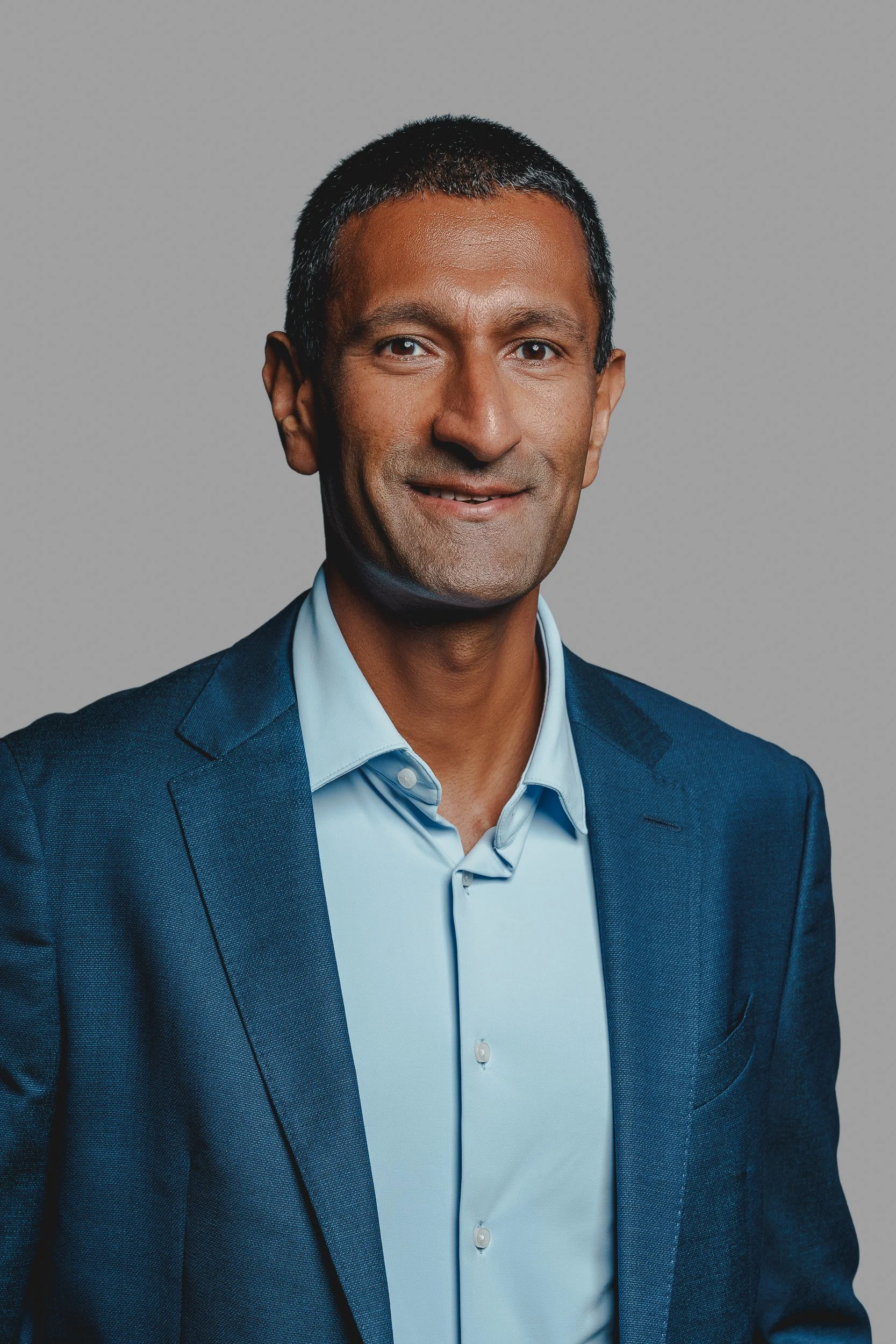 A professional portrait of a man wearing a blue suit and light blue shirt against a plain grey background.