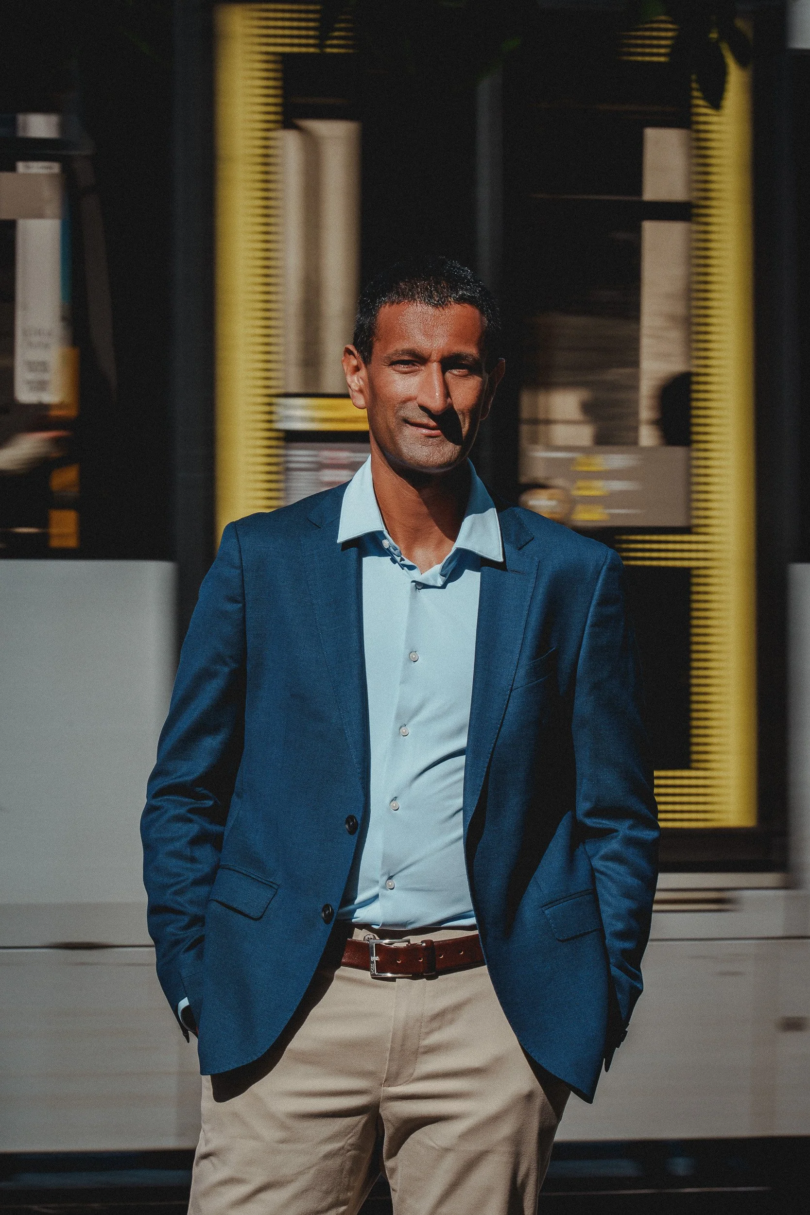 Dr Milan Mehta in a blue blazer, light blue shirt, and beige pants standing outdoors with a slight smile, with blurred background of a train passing behind him.