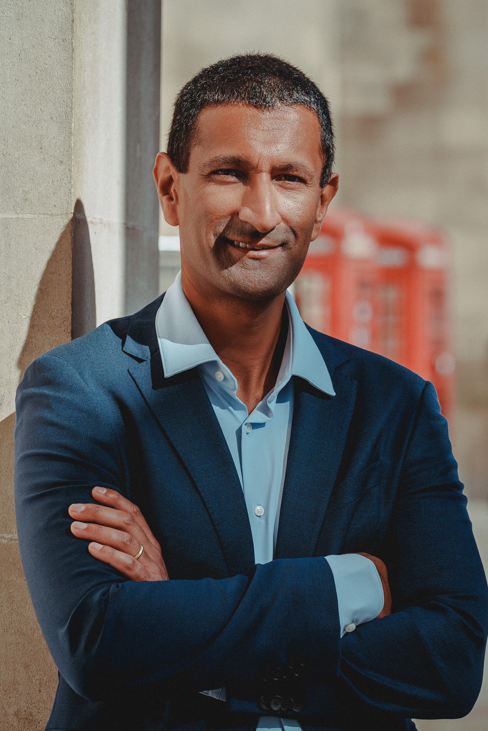 Dr Milan Mehta with short black hair wearing a blue suit with a light blue shirt, standing outdoors near a beige wall and smiling with his arms crossed.
