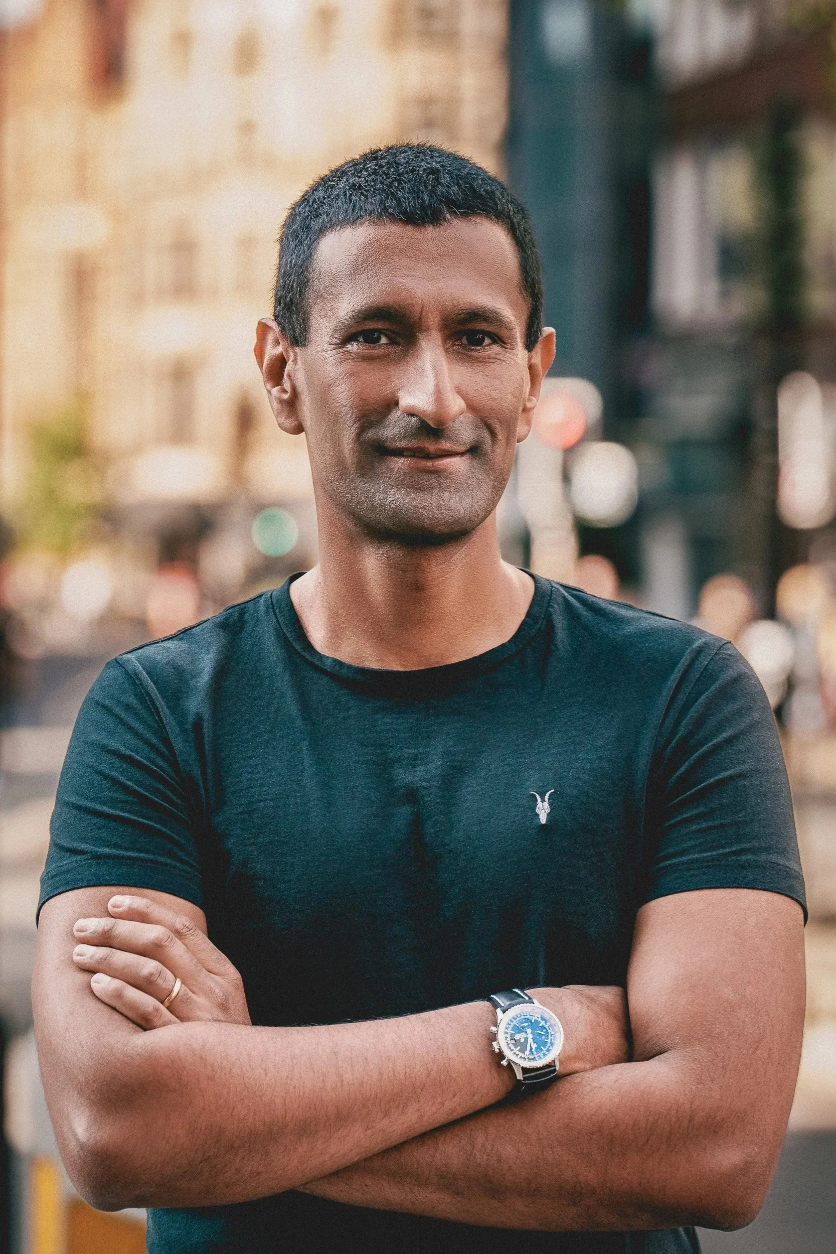 A man with short black hair and medium skin tone standing outdoors in front of blurred city buildings, crossing his arms, wearing a black T-shirt with a small emblem on the chest, a silver wristwatch, and a wedding band on his ring finger.
