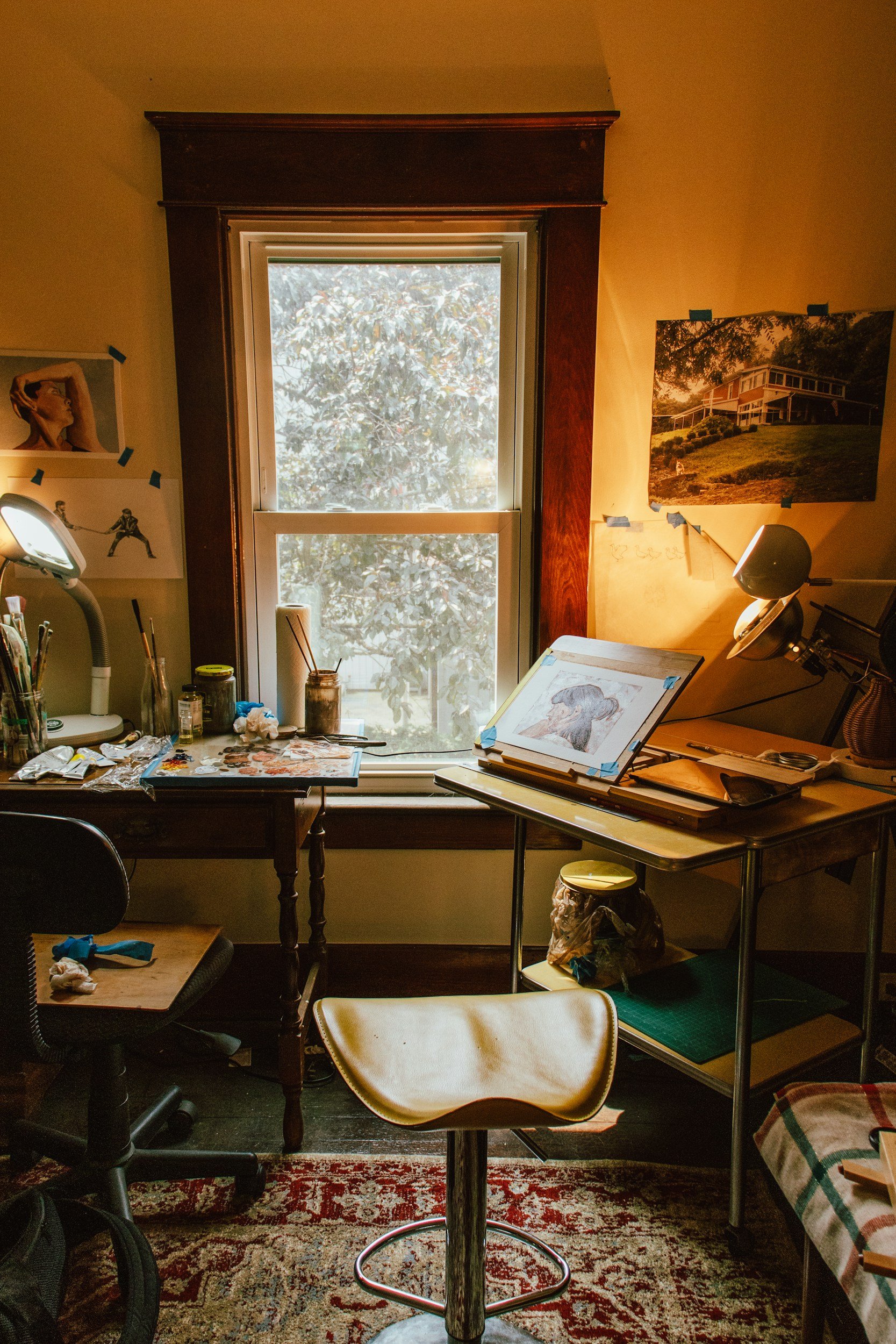 A cozy art studio with a window overlooking a leafy tree. The room has art supplies, sketches, and paintings on the walls, a table covered in art tools, and a vintage-style chair in front of a drawing easel.