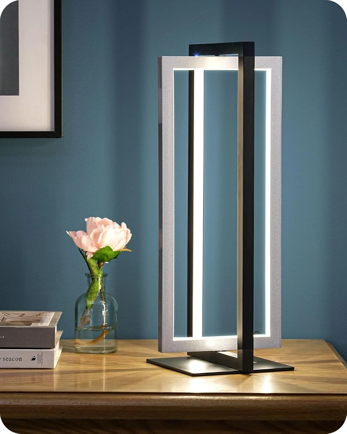 Modern table lamp with LED lights on a wooden desk. A pink flower in a glass vase and some books are also on the desk, with a blue wall background.
