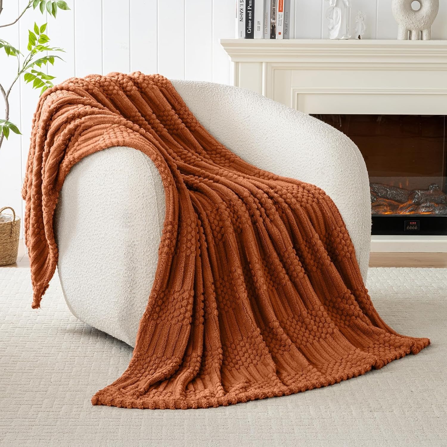 A white textured armchair in a living room with a rust-colored chunky knit blanket draped over it, a fireplace with a fire burning, a white mantle with decorative objects, a bookshelf with books, a green plant, and a neutral-colored rug.