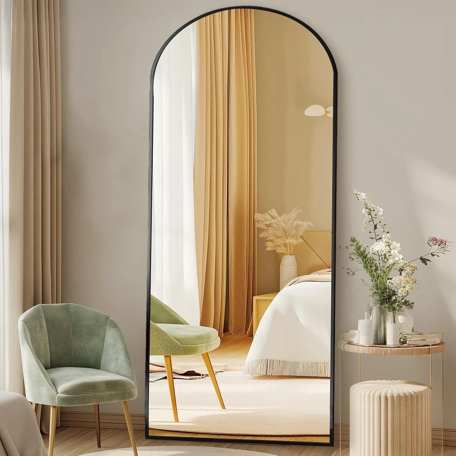 A large, arched mirror reflects a cozy bedroom with a beige nightstand, a vase with dried pampas grass, and a bed with white bedding. Next to the mirror is a side table with a vase of flowers.