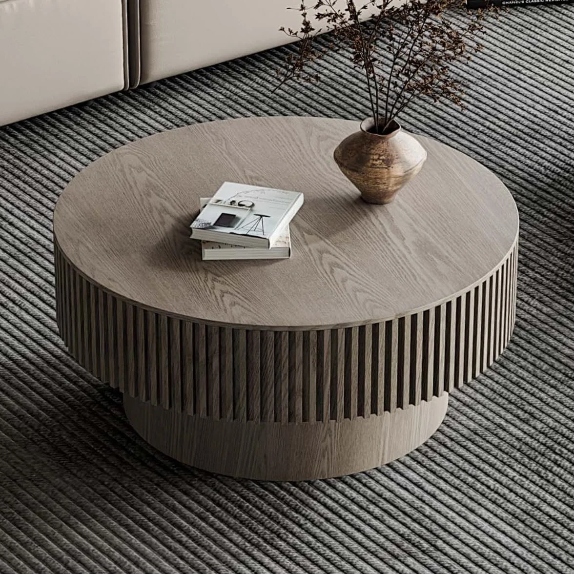 Round wooden coffee table with vertical slats on the sides, placed on a textured striped rug. The table has a ceramic vase with dried branches and a few stacked books with a key and glasses on top.