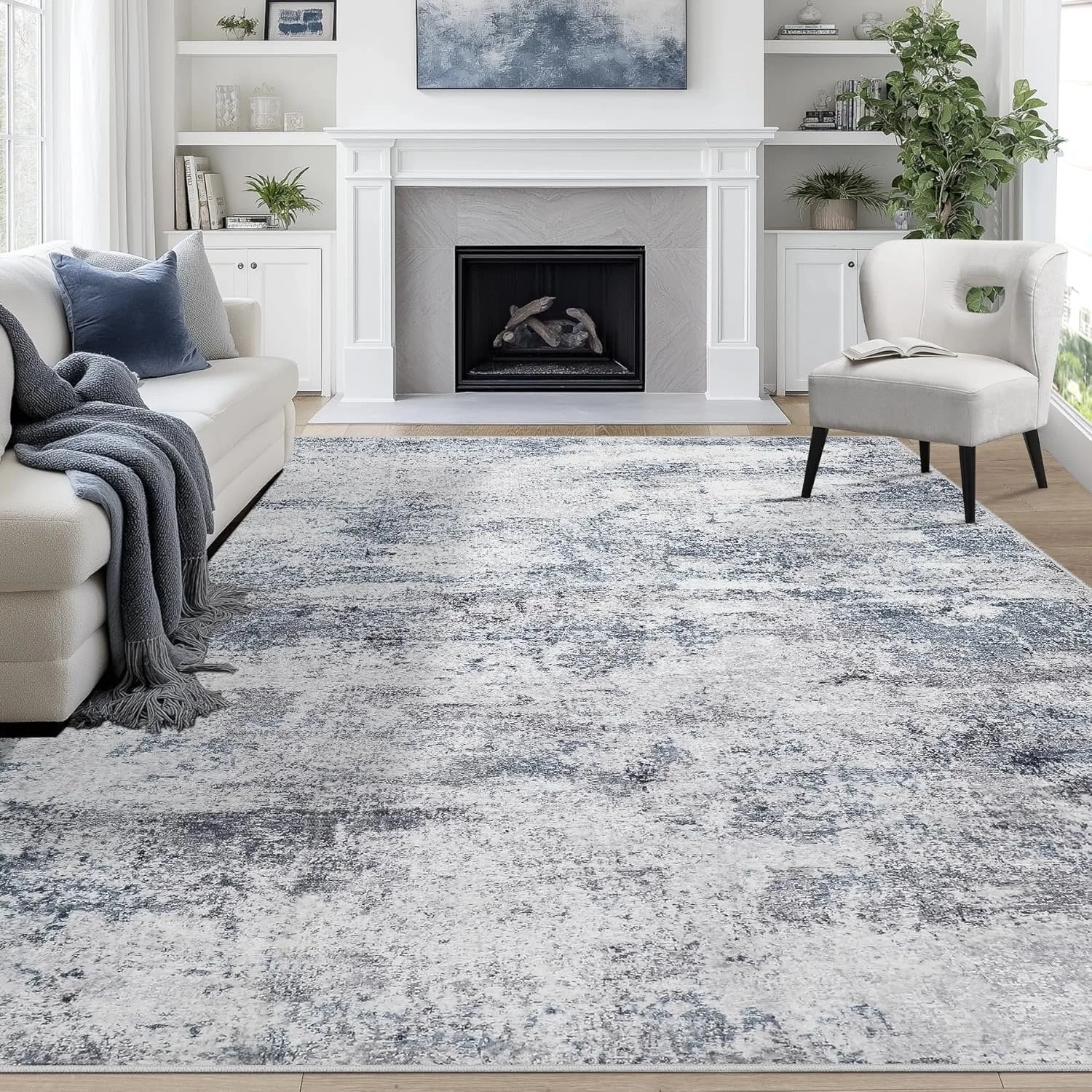 Living room with white sofa, gray and blue throw pillows, a gray throw blanket, a white armchair with an open book, a patterned area rug, a white fireplace with plants on both sides, built-in bookshelves, and framed artwork above the fireplace, with natural light coming from large windows.