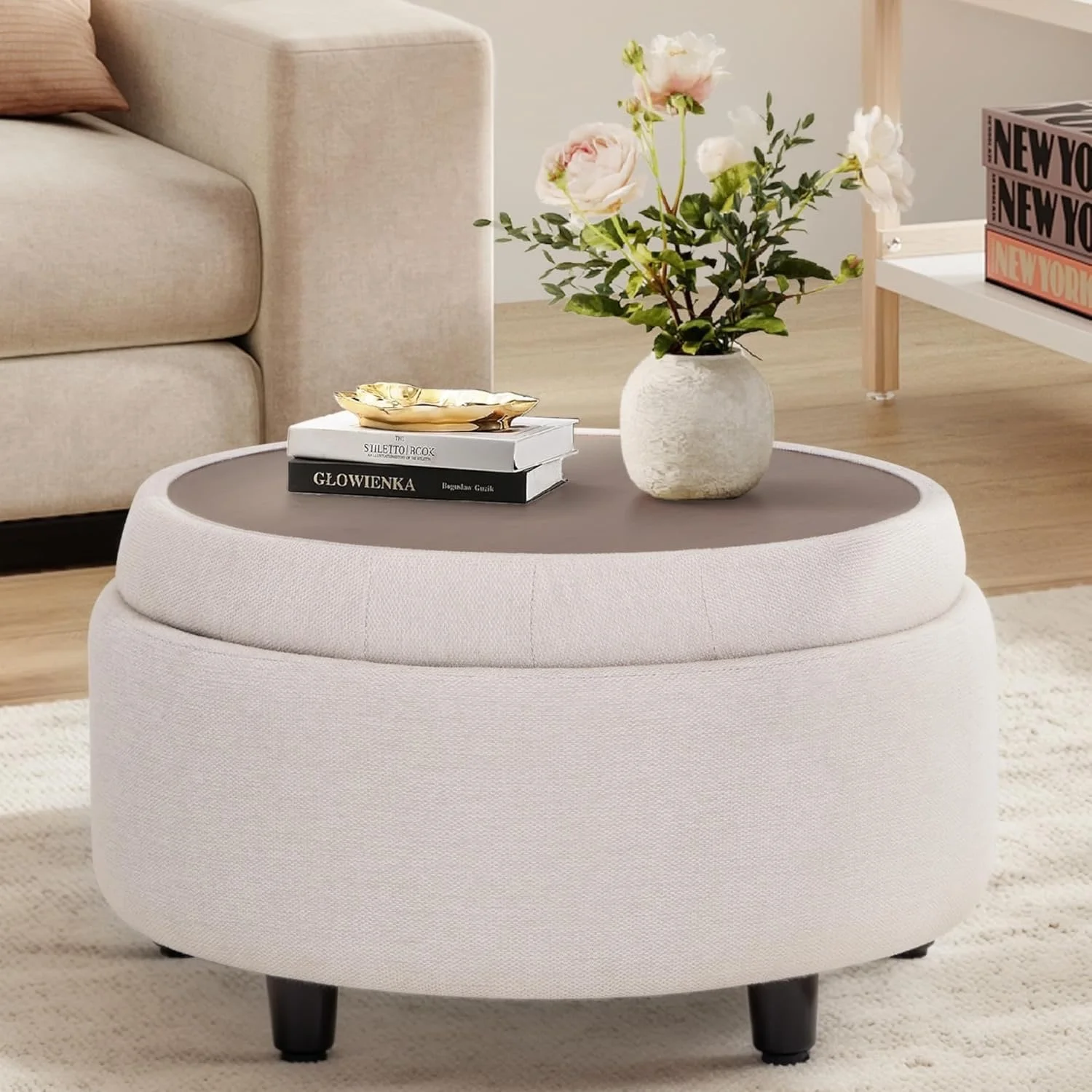 A round, light-colored upholstered ottoman with black legs, topped with a white book, a smaller black book, a gold dish, and a white vase with pink and white flowers, in a cozy, modern living room.