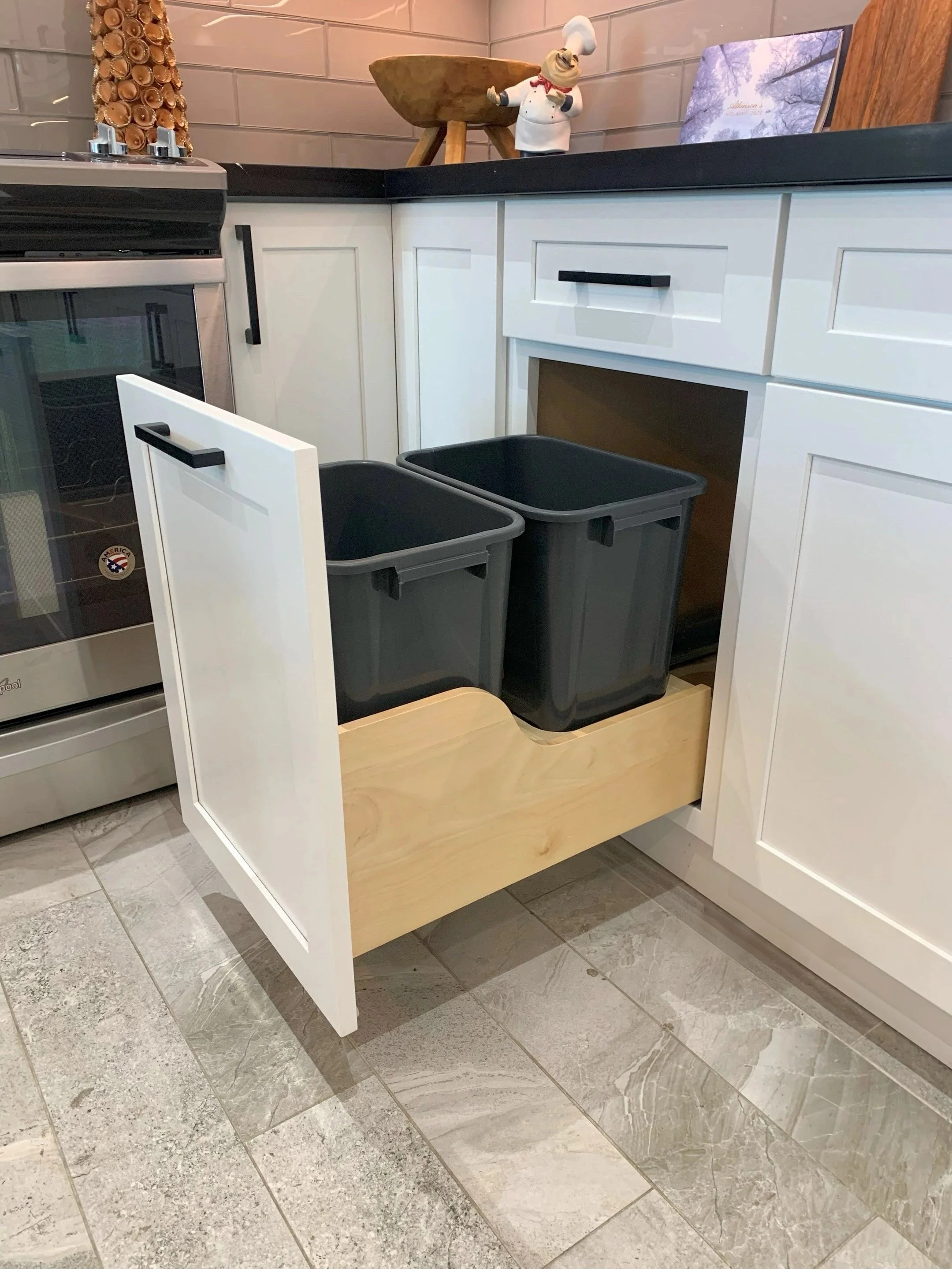 Open kitchen cabinet drawer with two black trash bins inside, white cabinets, black countertop, decorative items including a chef figurine, a wooden bowl, and a decorated cone-shaped item in the background. Floor is tiled with grayish stone tiles.
