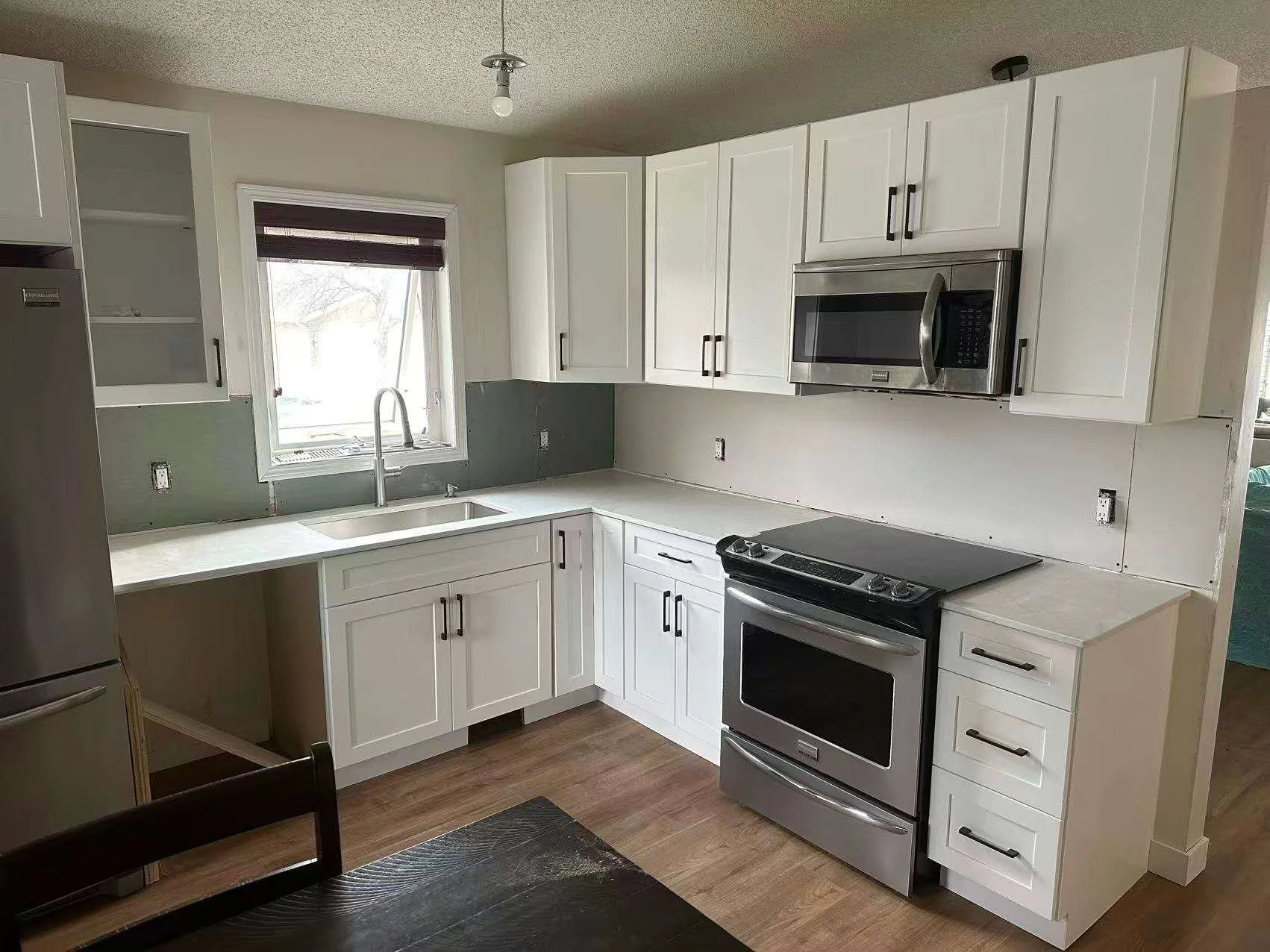 Kitchen with white cabinets, stainless steel microwave, oven, and refrigerator, black handles, under renovation, with open wall outlets and a window above the sink.
