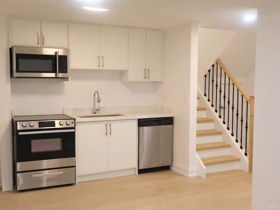 Kitchen with white cabinets, stainless steel microwave, stove, dishwasher, and a sink, next to a staircase with wooden steps and a black metal railing.