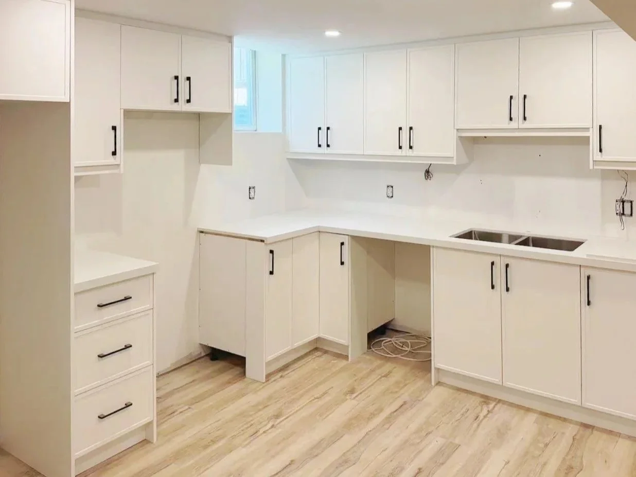 Empty kitchen with white cabinets and drawers, a partially installed double sink, light wood flooring, and a small window.