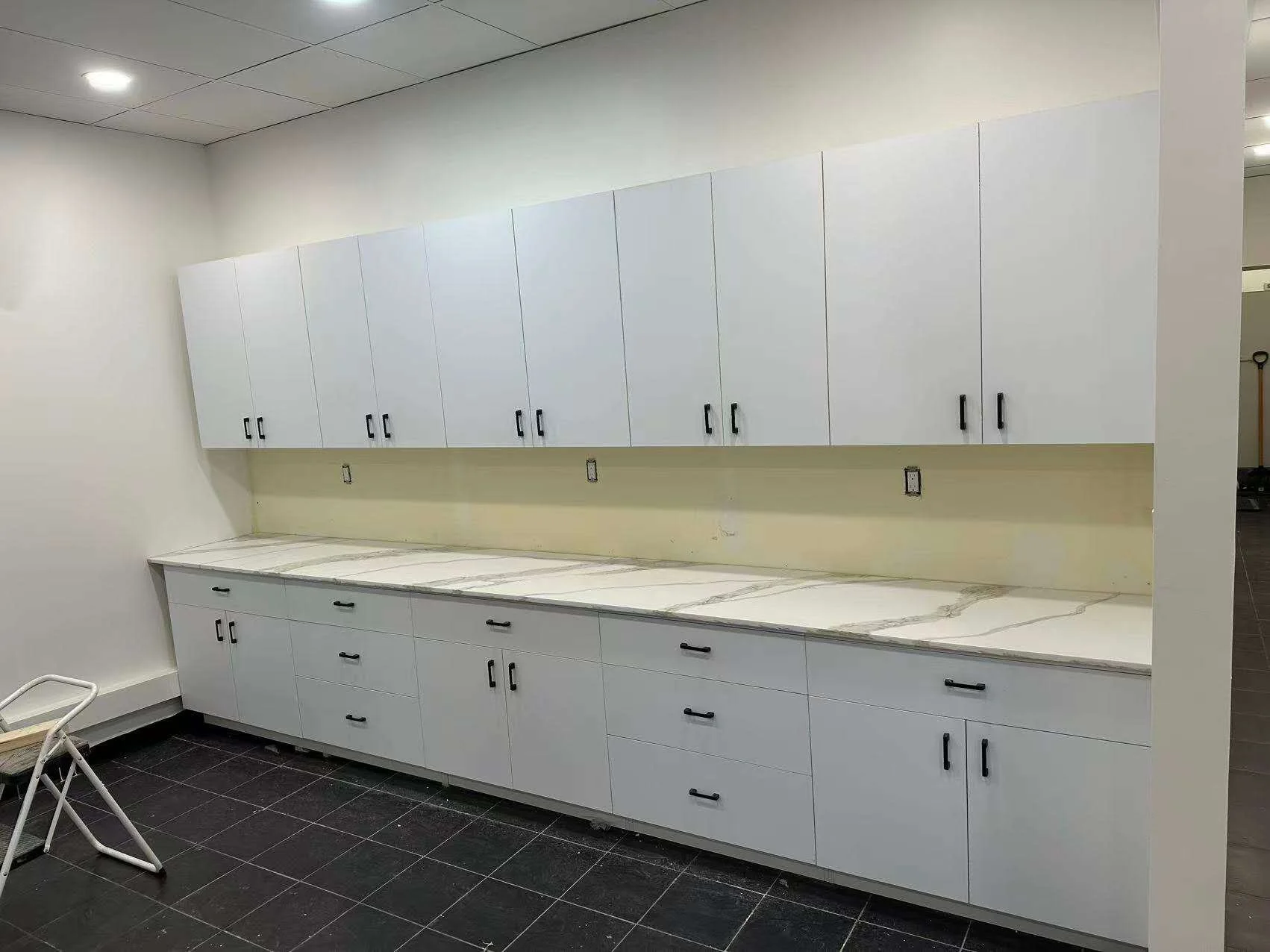 Empty kitchen or utility room with white cabinets, marble countertop, and black handles. The room has a tile floor, electric outlets on the wall, and a ceiling with paneling. A small white step stool is in the foreground.