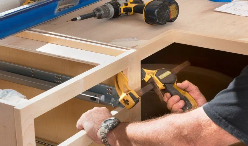 A person working on a woodworking project, using a cordless drill and a yellow cordless screwdriver to assemble a wooden frame at a workbench.