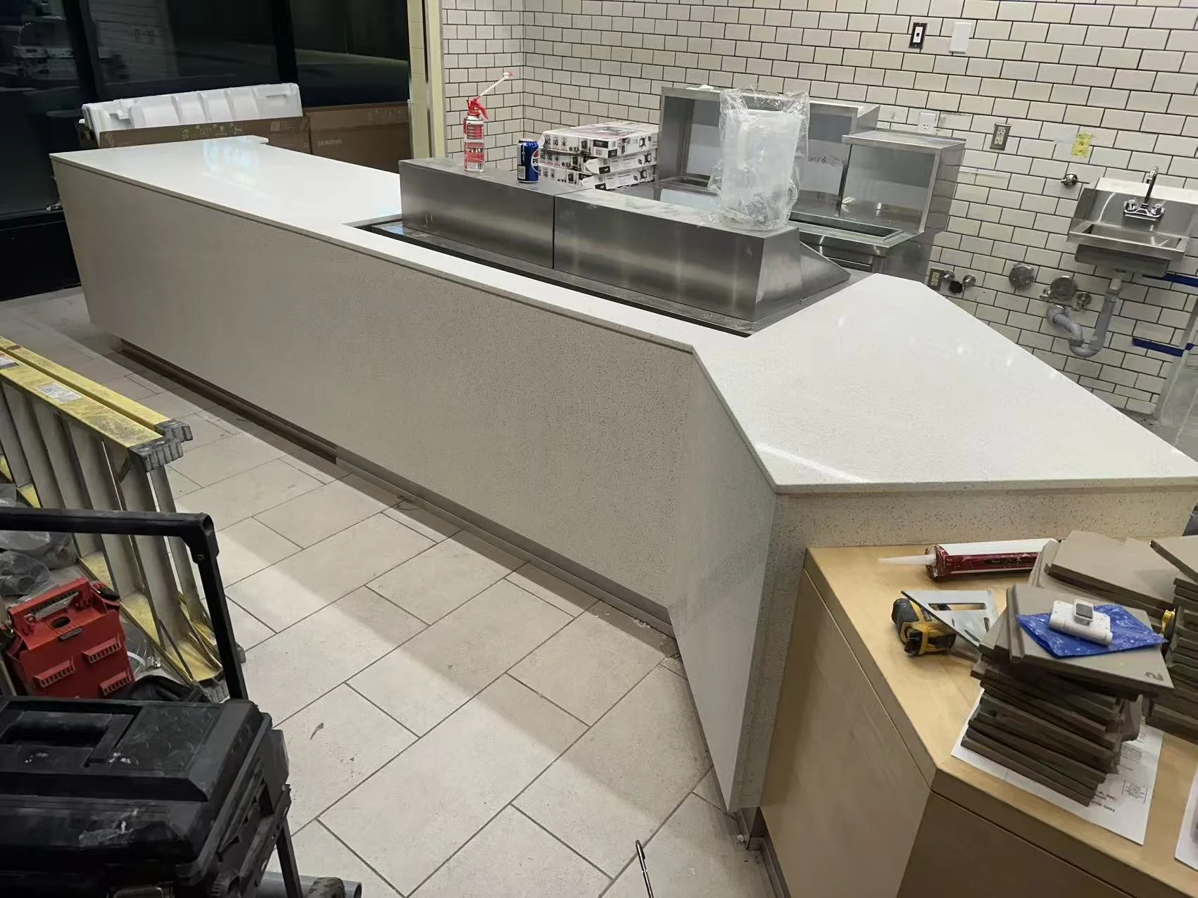 Construction scene inside a commercial space with a long white countertop, construction tools, stacked materials, and stacks of paperwork. The background has brick walls and plumbing fixtures.