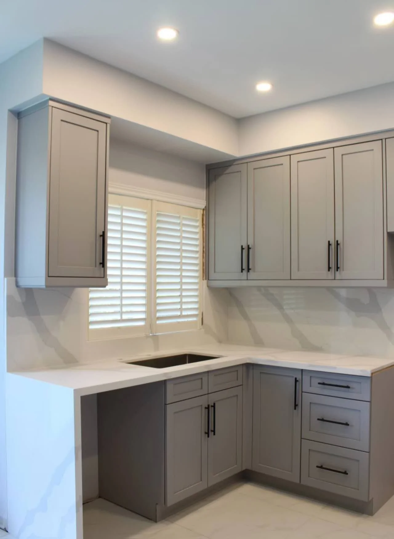Kitchen with gray upper and lower cabinets, white countertops, a window with white shutters, and recessed ceiling lights.