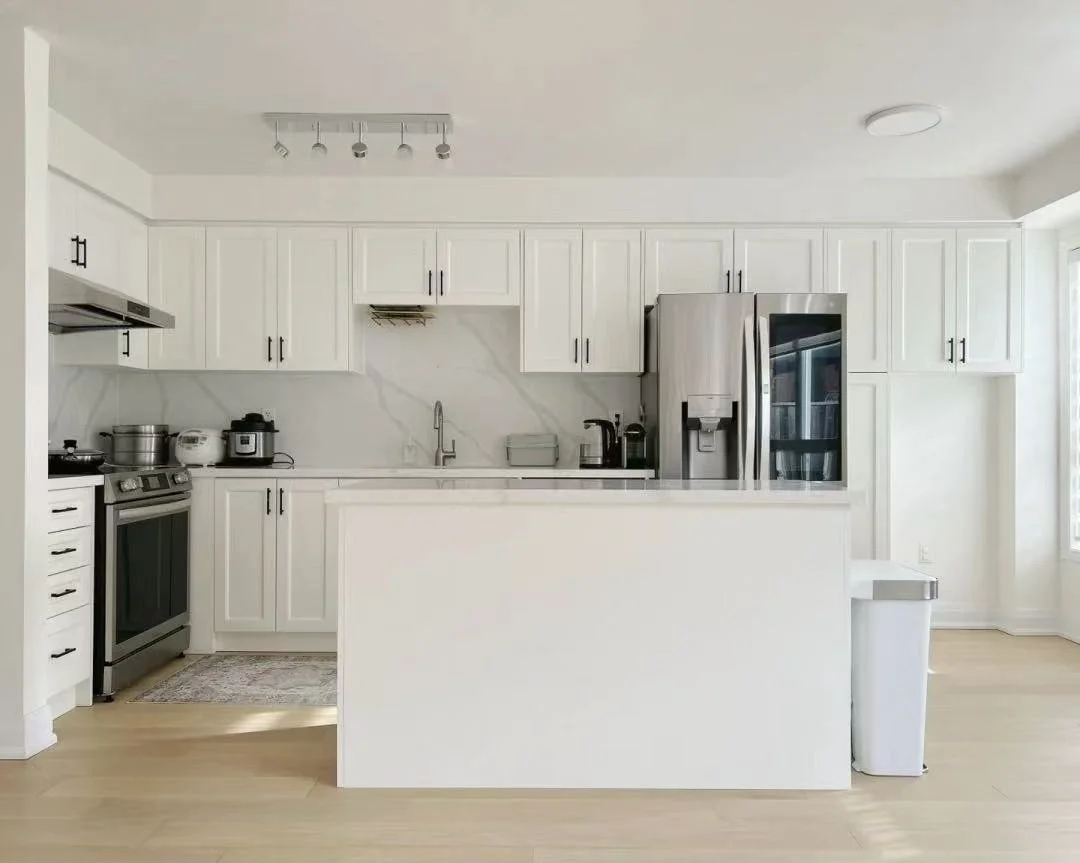 A modern white kitchen with stainless steel appliances, including a refrigerator, oven, and microwave, and a marble backsplash.
