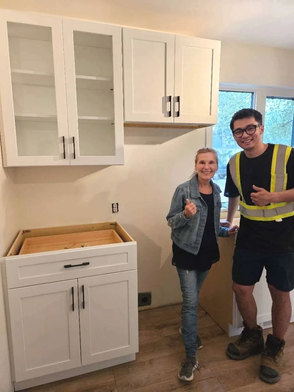 Two people smiling in a kitchen with white cabinets, one in a denim jacket and the other in a construction vest, standing near a countertop and a window.