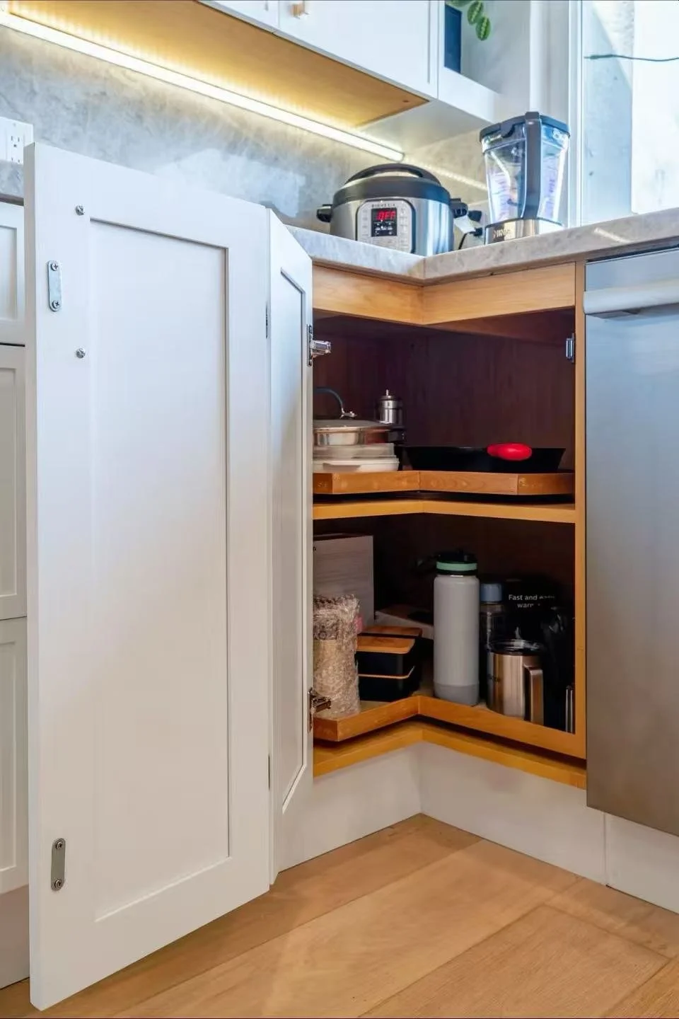 Kitchen corner with open cabinet containing containers, a water bottle, and kitchen appliances, with countertop appliances like a rice cooker and blender visible above.
