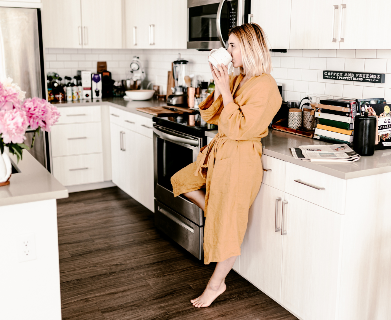 A woman in a tan bathrobe drinking from a mug in a modern kitchen with white cabinets, a stove, a microwave, black and white appliances, and pink flowers on the counter.