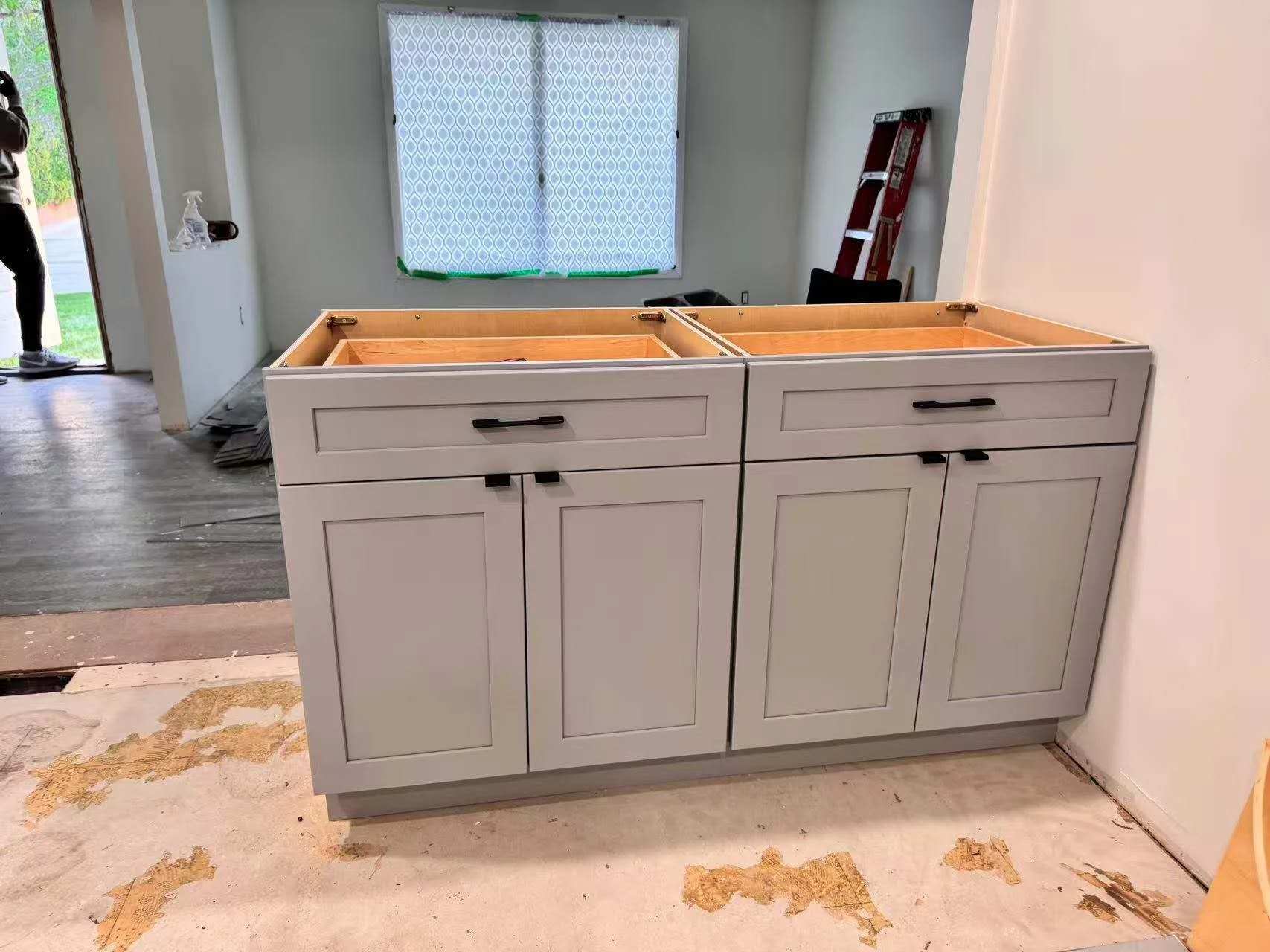 A partially assembled kitchen cabinet with two drawers on top and two doors below, in a room under construction with a window, ladder, and construction materials.