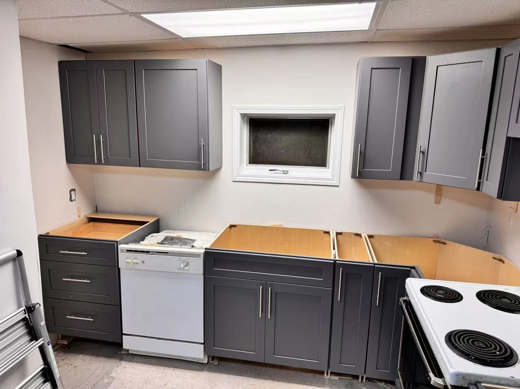 Kitchen with grey cabinets, a window, a white dishwasher, and a white stove with coil burners. The cabinets are being installed or renovated, and some cabinet doors are missing. There is a ladder on the left side.