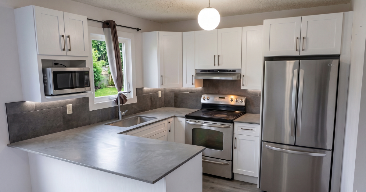Kitchen with white cabinets, stainless steel refrigerator, microwave, oven, and gray countertops. A window with a dark curtain, overhead light, and gray backsplash.