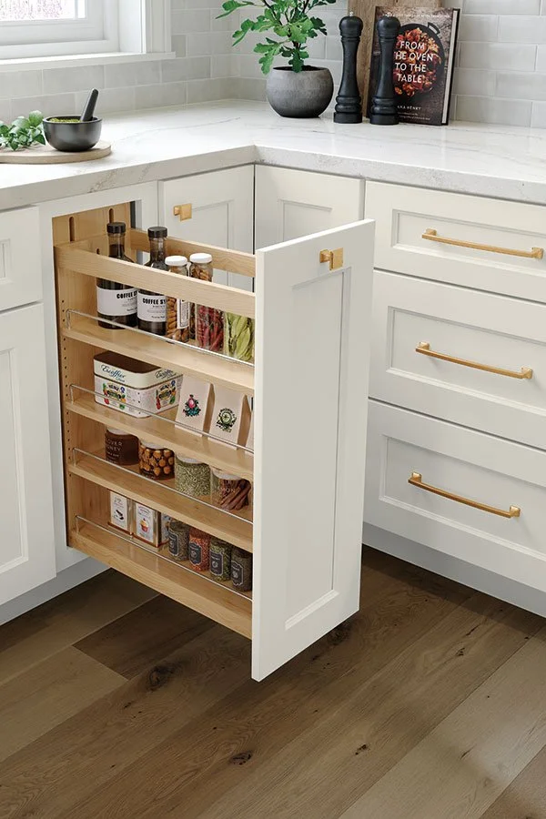 Open kitchen cabinet with pull-out spice rack containing jars of spices and seasonings, white cabinetry with gold handles, and a light-colored countertop with decorative items on it.