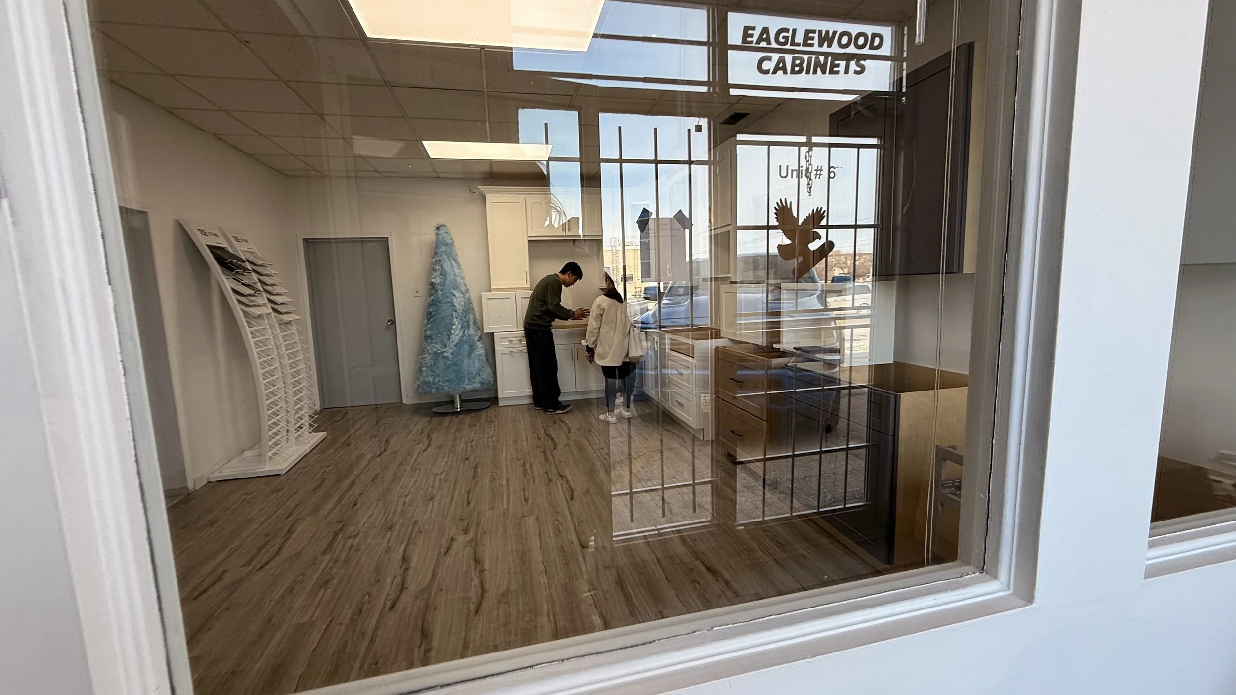 Interior view of a showroom or office displaying white cabinets, a blue Christmas tree, and two people examining the cabinets through a glass window with a sign that reads 'Eaglewood Cabinets' and 'Unit #6'.