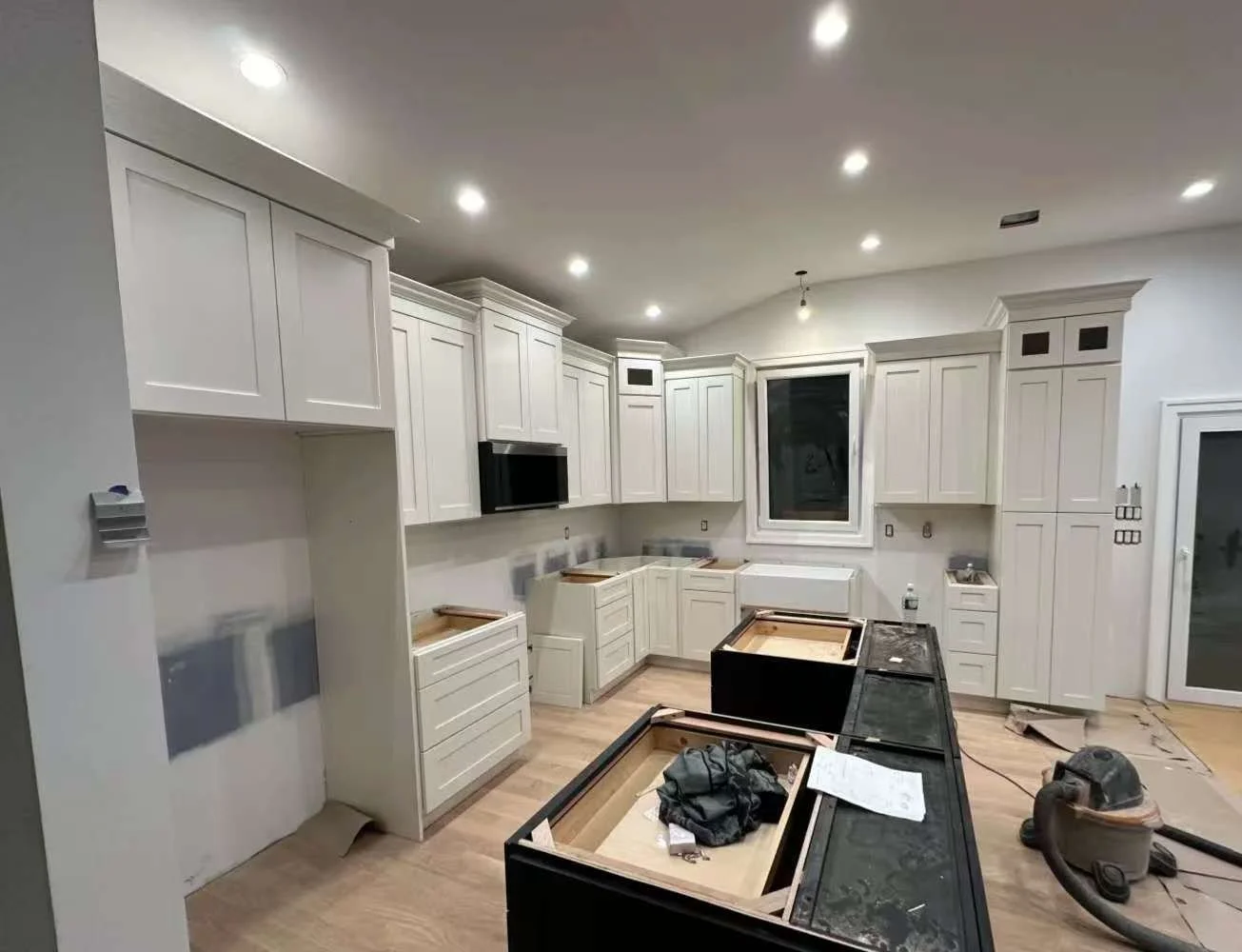 Kitchen cabinets under construction with black countertops, a window, and a nearby vacuum cleaner on the floor.