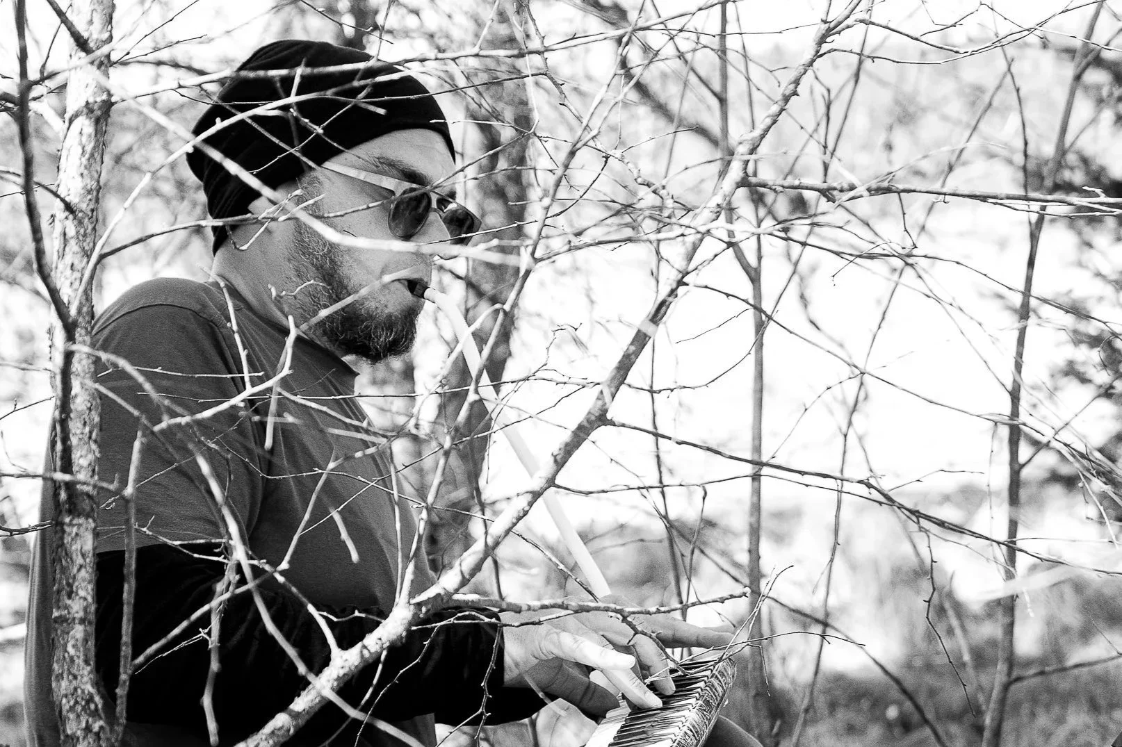 A man wearing sunglasses, a headwrap, and a t-shirt is playing a keyboard outdoors behind leafless trees.