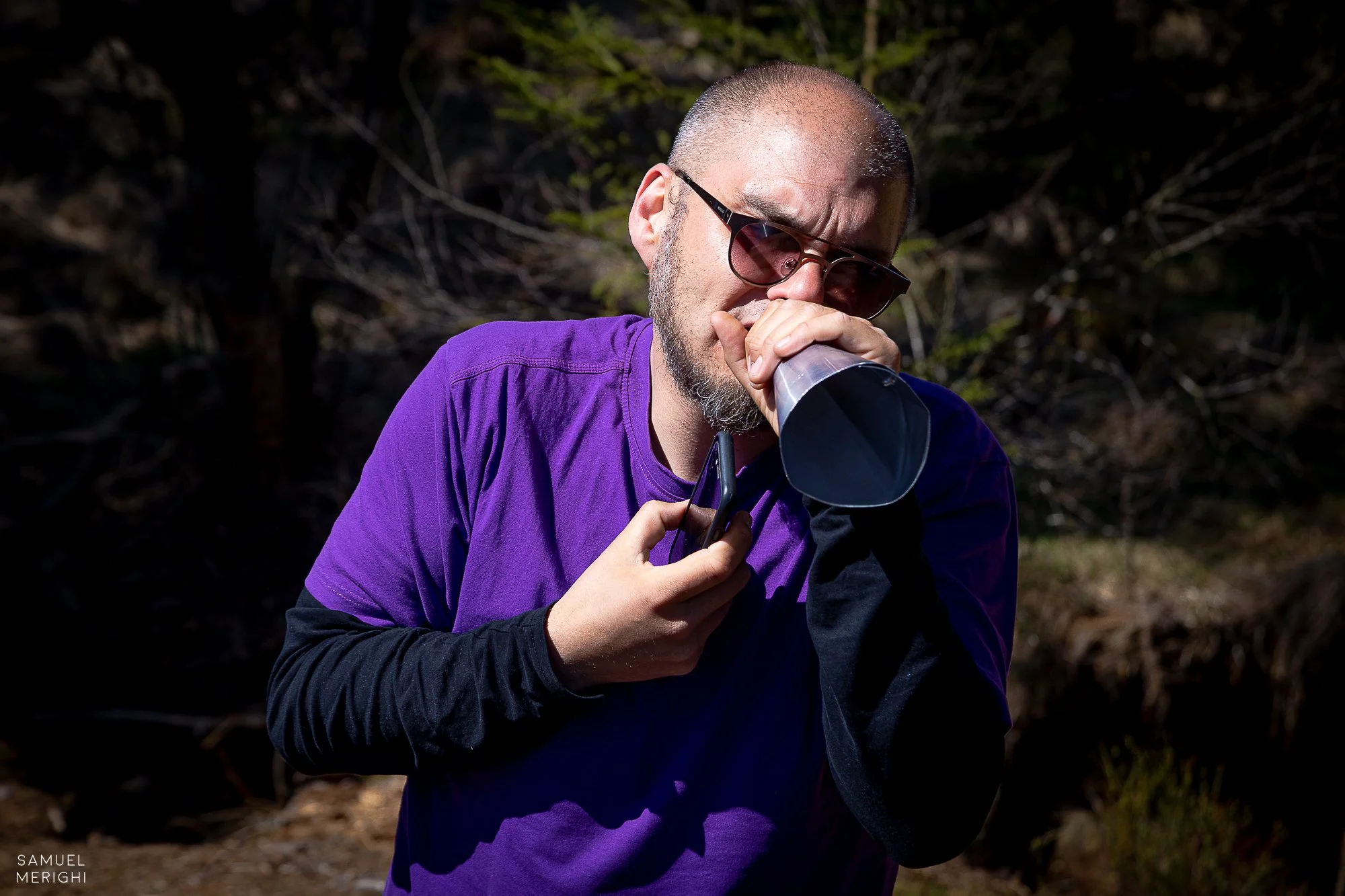 A man with a shaved head and beard wearing sunglasses and a purple jacket is drinking from a cup outdoors.