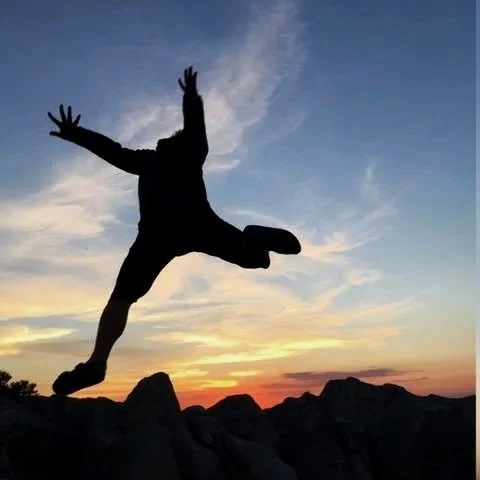 Silhouette of a person jumping with arms and legs spread wide against a sunset sky over rocky terrain.