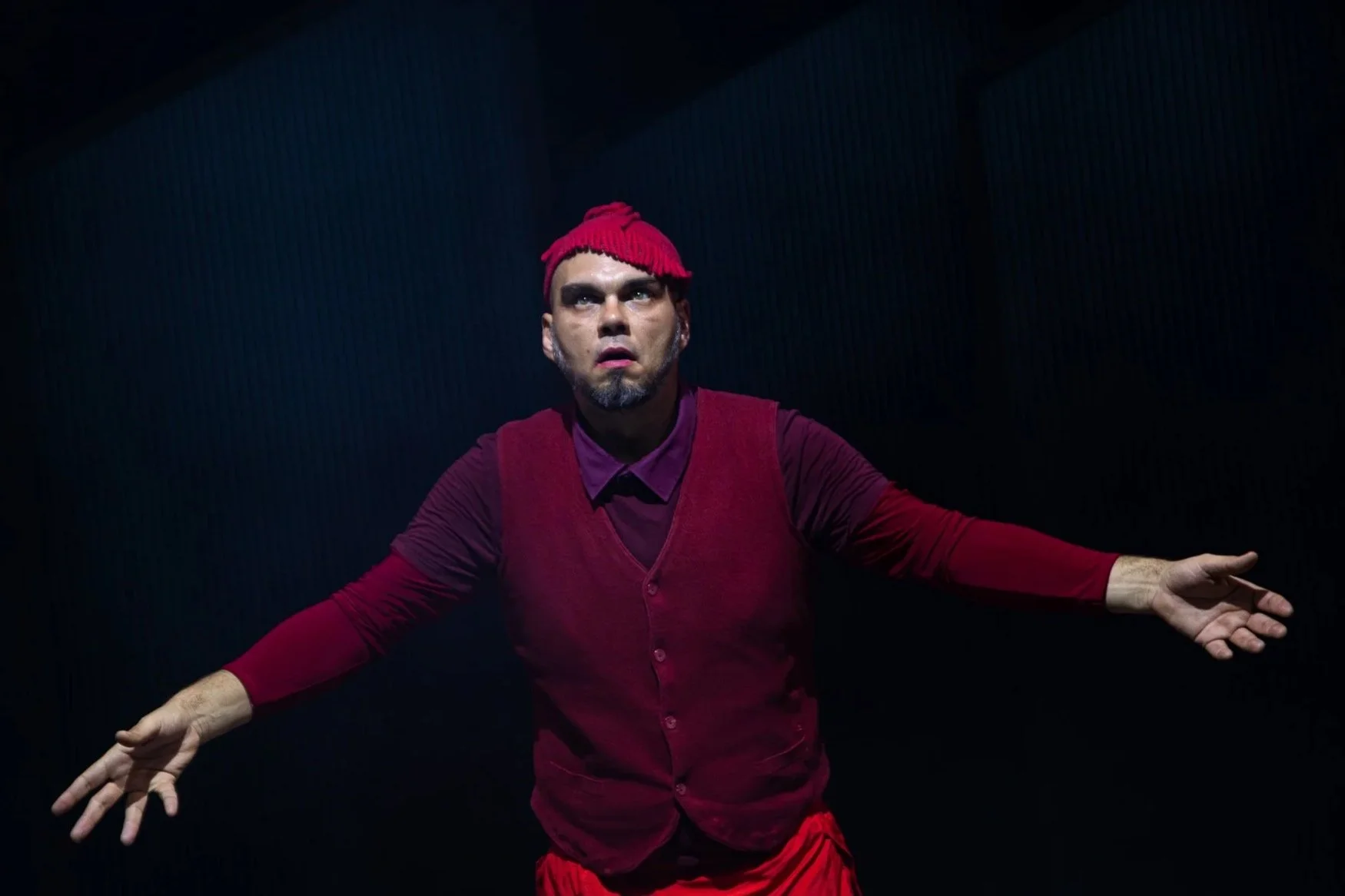 A person with a short gray beard, wearing a red knit hat, maroon vest, and long-sleeve shirt, standing against a dark background with arms extended.