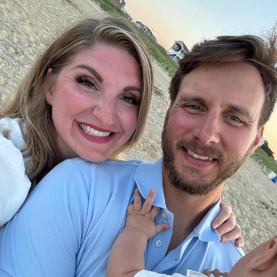 A smiling woman and man taking a selfie on the beach at sunset. The woman has blonde hair and is wearing a white top, while the man has brown hair and a beard, wearing a light blue collared shirt. The woman has her arm around the man's shoulder, and the man's arm is extended to hold the camera. In the background, there are beach houses and some trees.
