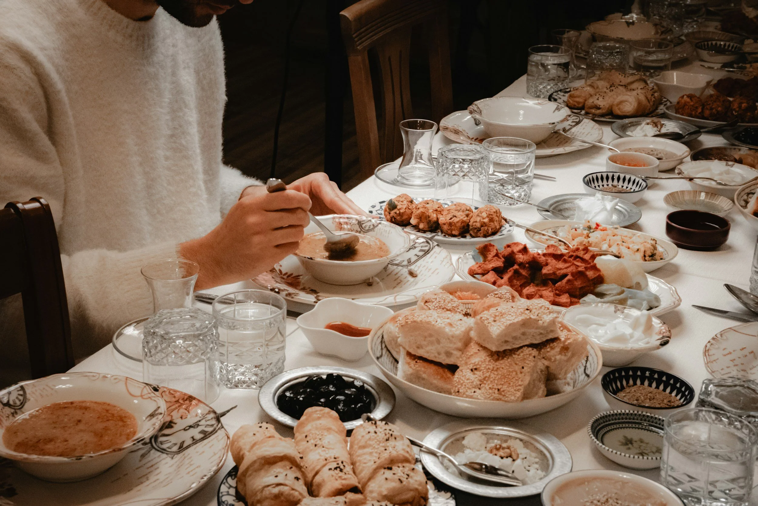 A person in a white sweater is serving food at a large, well-set dining table filled with various dishes, bowls, and glasses, indicating a meal or celebration.