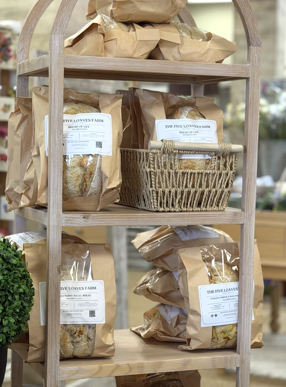 Wooden display shelf with paper bags containing bread from Five Loaves Farm, labeled with different bread varieties such as sourdough, bagel bread, and Italian cheese or banana bread.